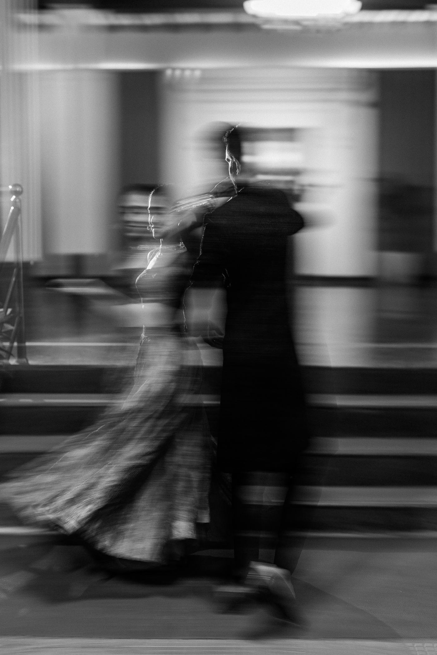 Motion-blurred black and white photograph shows silhouettes of dancers in flowing dresses performing on a stage.
