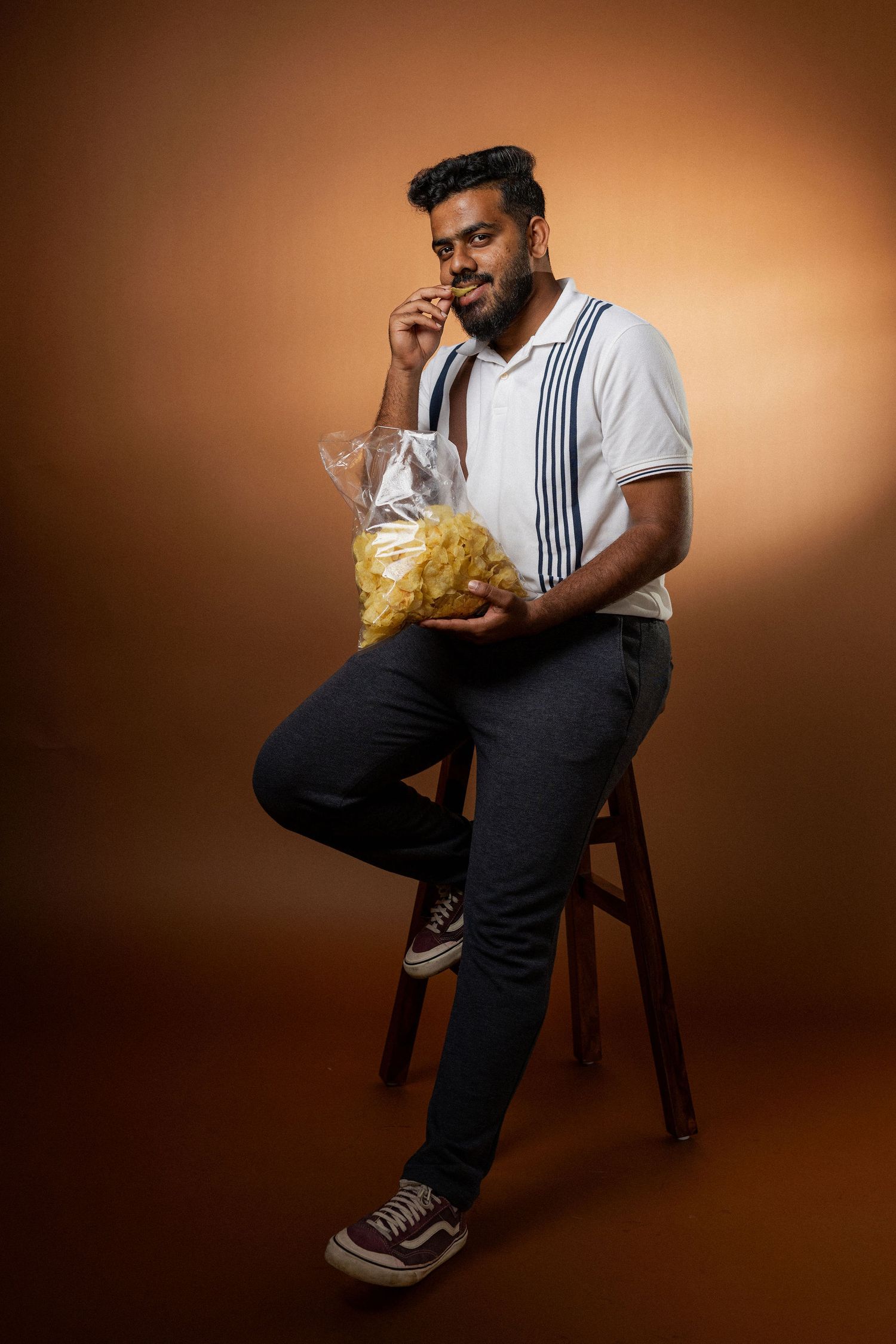 A person in suspenders sits on a wooden stool eating potato chips from a clear bag against an orange-brown background.