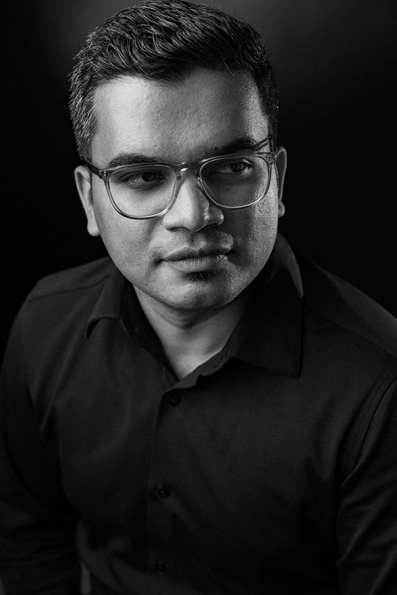 Black and white professional portrait of a person wearing glasses and a dark polo shirt against a dark background.