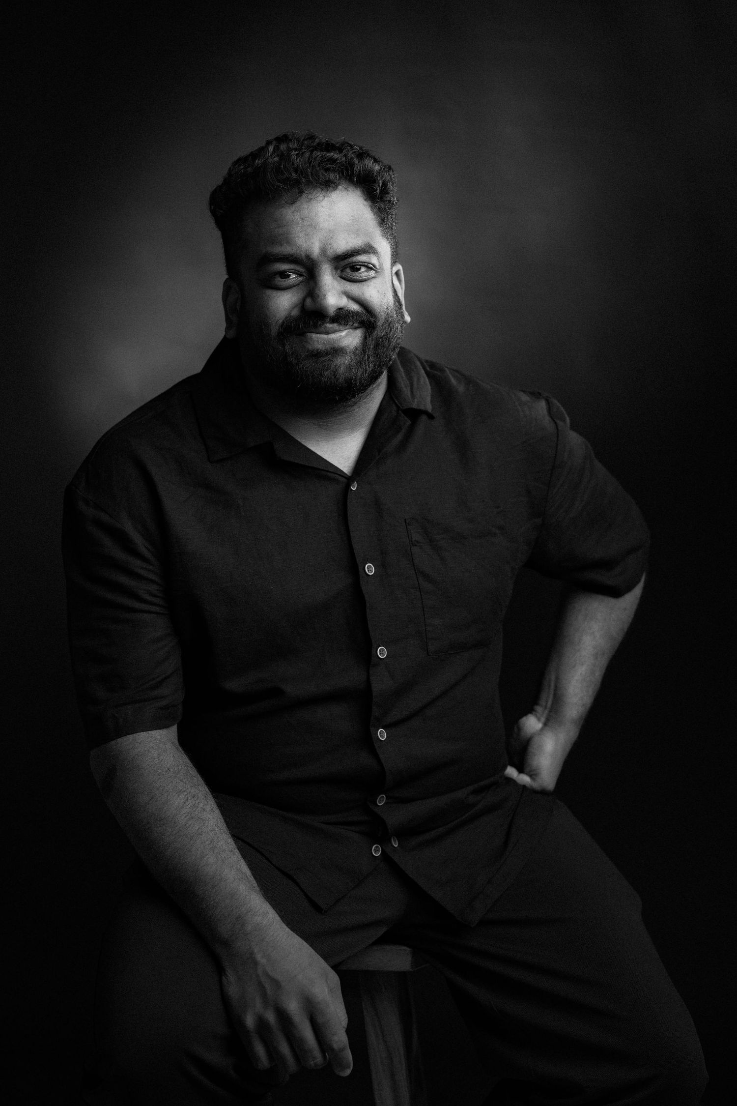 Black and white portrait of a seated person in dark clothing with a warm smile against a dark background.