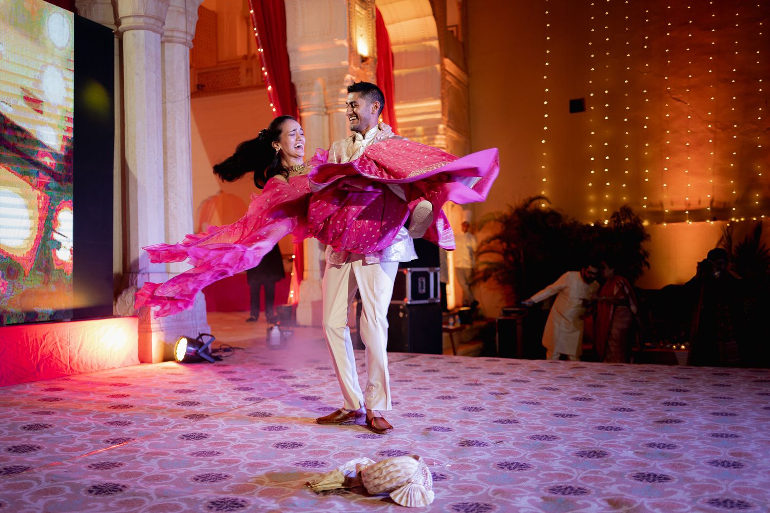 Couple dancing together at a wedding reception with pink lighting and string lights creating a romantic atmosphere.