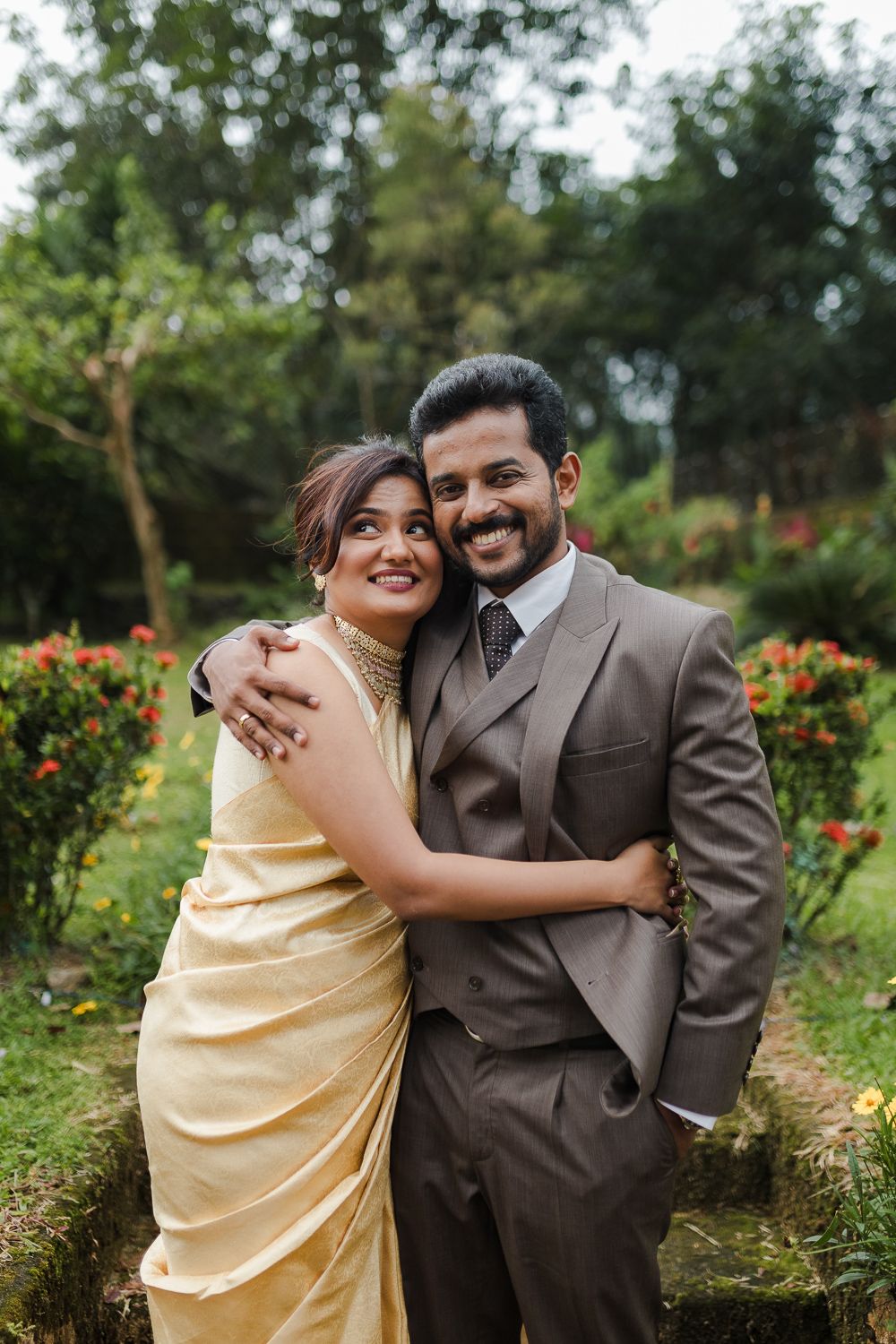 A couple embraces in a garden with blooming flowers, wearing formal attire of brown suit and cream dress.