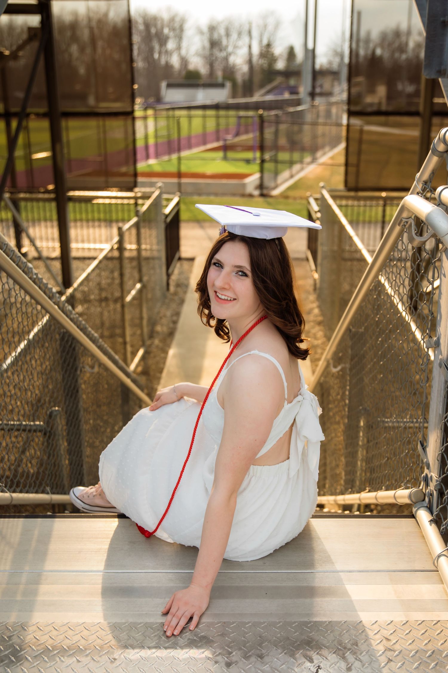 A smiling young woman in a white dress and graduation cap sits on metal bleacher steps at a sports field.