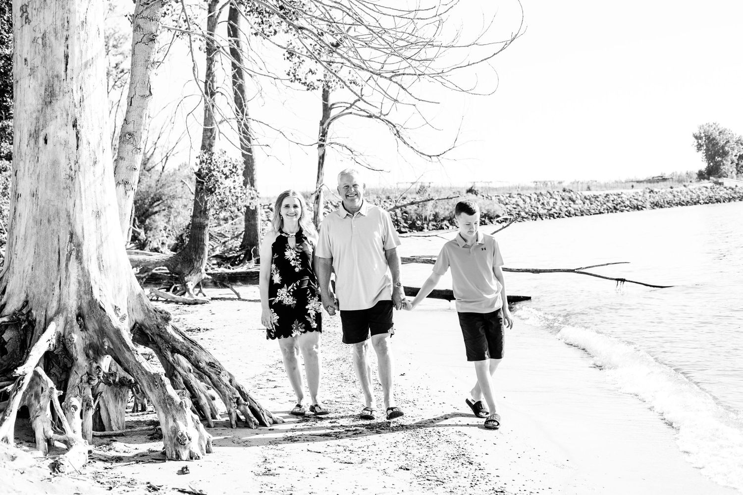 Three people stand together near large tree roots at a beach in black and white.