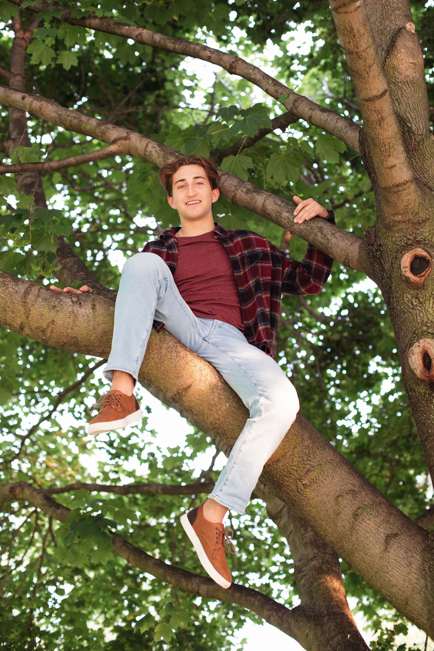 Playful portrait sitting in tree wearing plaid shirt and light colored pants.