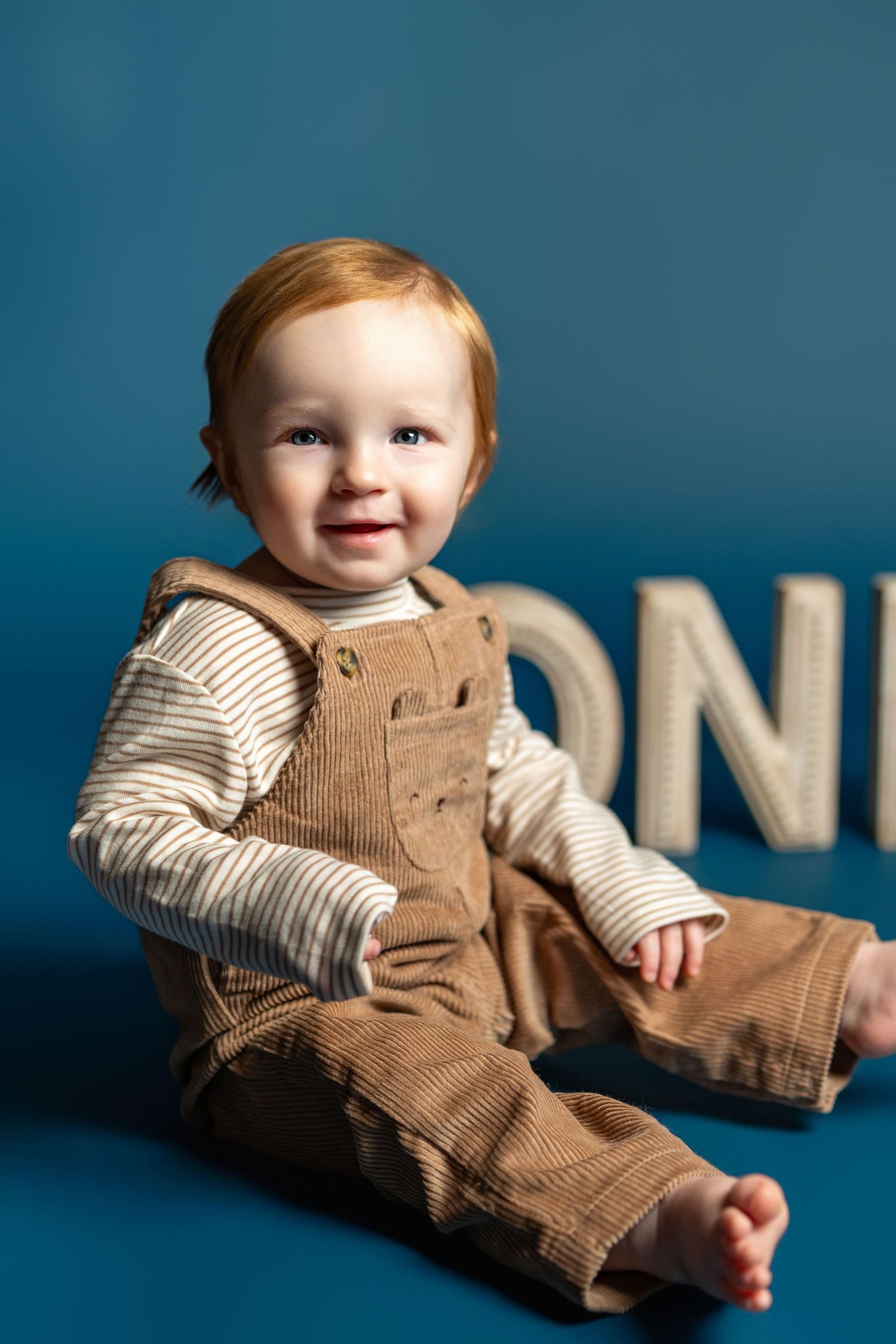Baby wearing brown corduroy overalls and striped shirt sits on blue backdrop with white letters in studio setting.