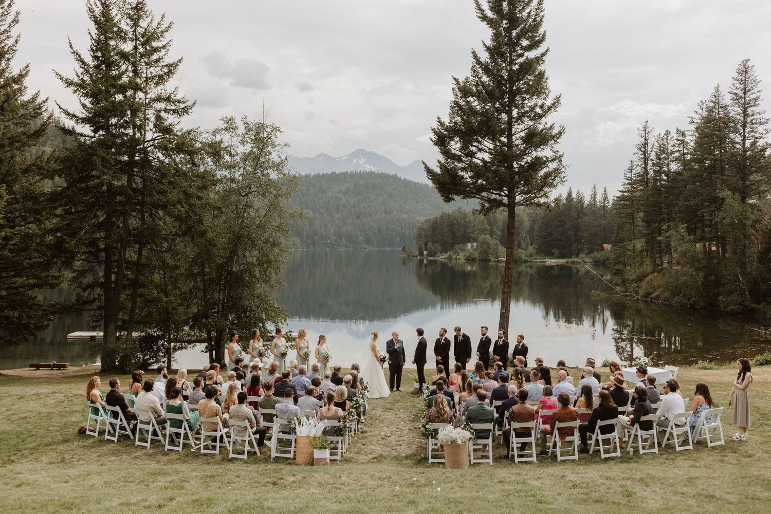 A panoramic view of an outdoor wedding ceremony set against a scenic mountain lake backdrop with evergreen trees.