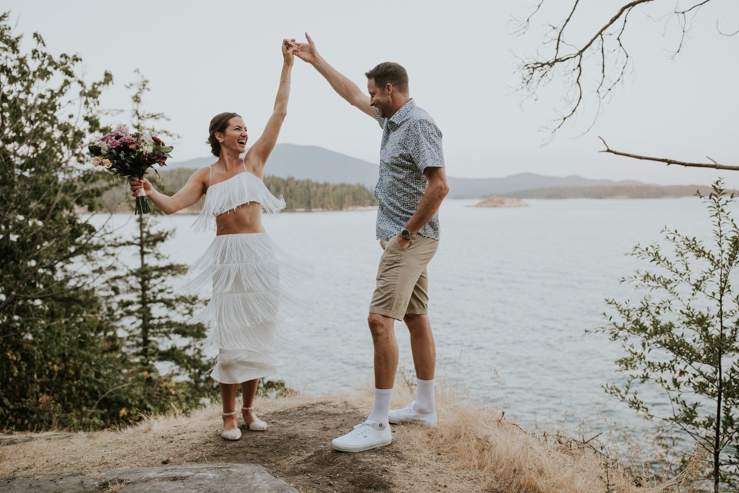 A couple celebrates their wedding day dancing on a cliff overlooking a scenic lake surrounded by trees and mountains.