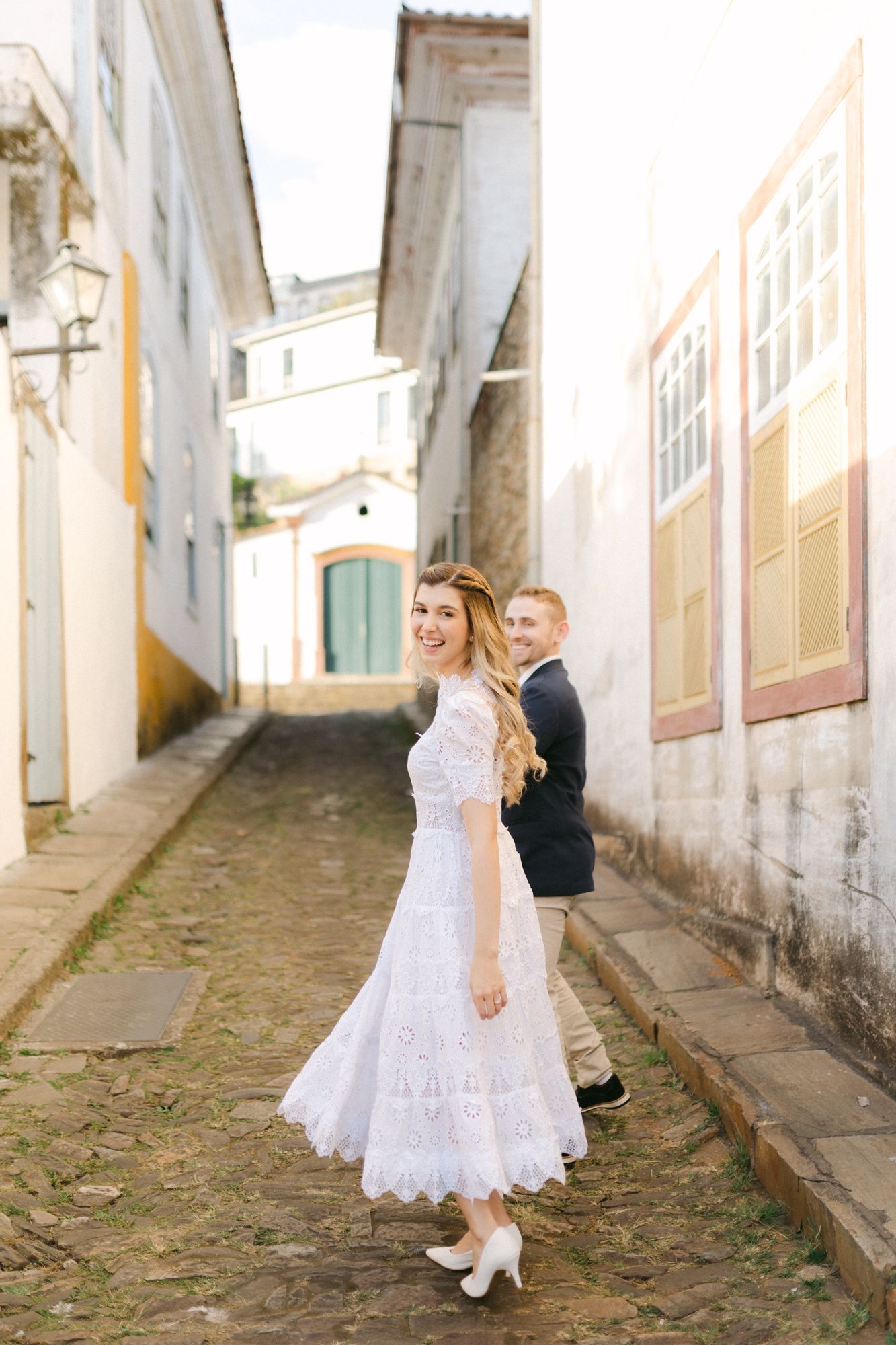 Couple posing back-to-back in white dress and dark outfit on quaint cobblestone street.