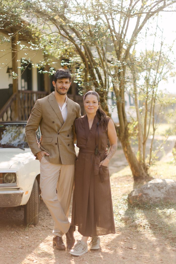 A couple in vintage-style brown attire poses outdoors together in a sunlit wooded setting.