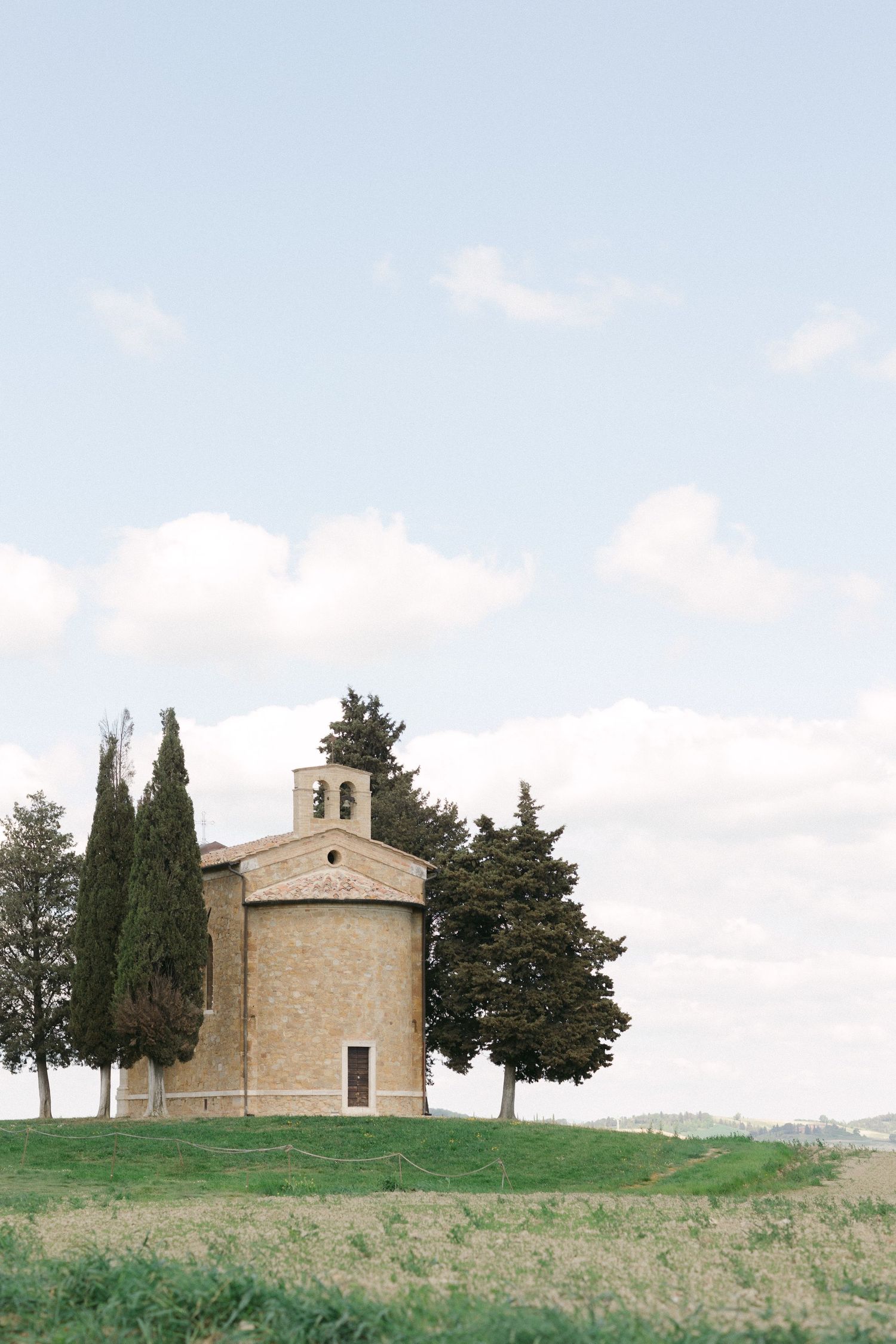 Small rustic chapel with bell tower stands alone on grassy hill surrounded by cypress trees in Tuscan countryside.