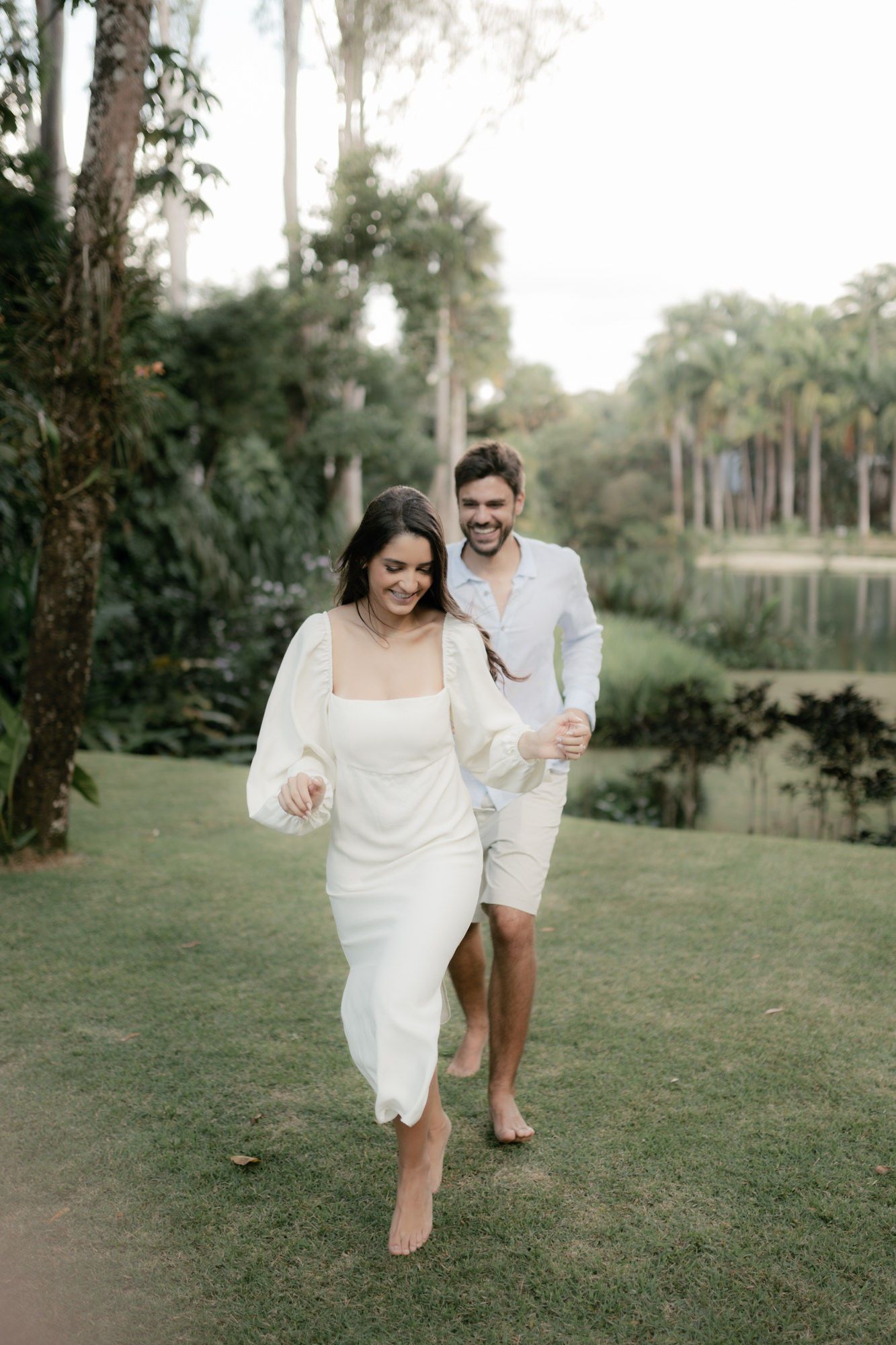 A couple runs playfully through a lush garden setting with palm trees and a lake.