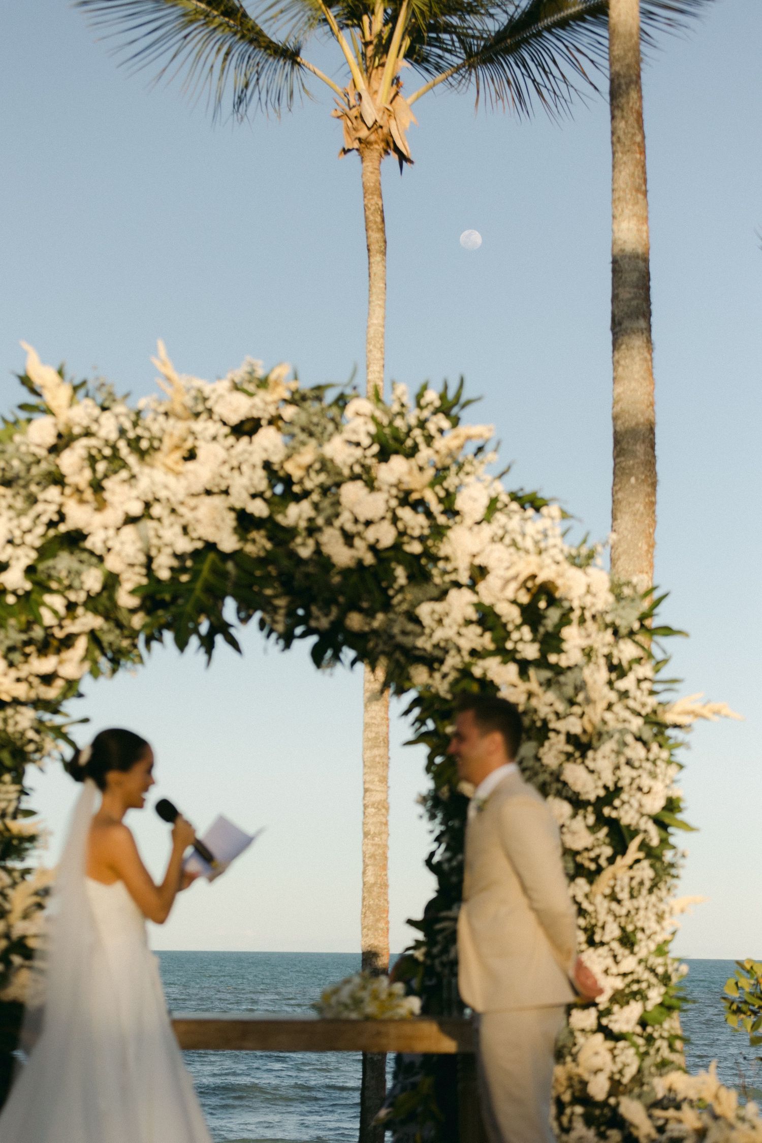 A romantic seaside wedding ceremony takes place under a white floral arch with palm trees and ocean views at sunset.