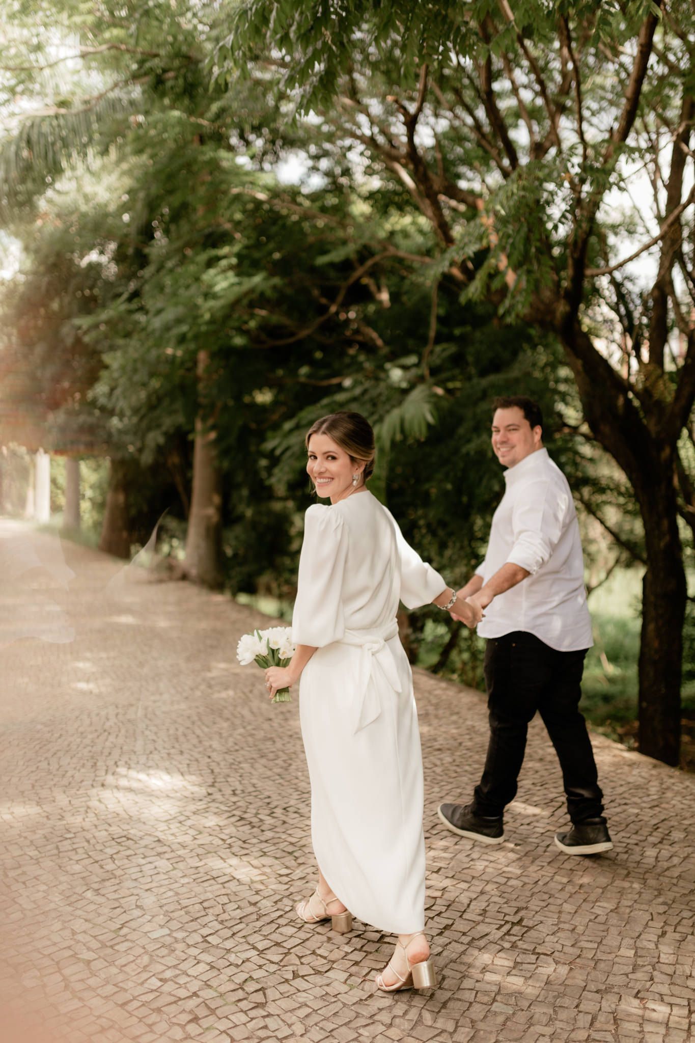 Couple in white attire walking on cobblestone path surrounded by lush greenery.