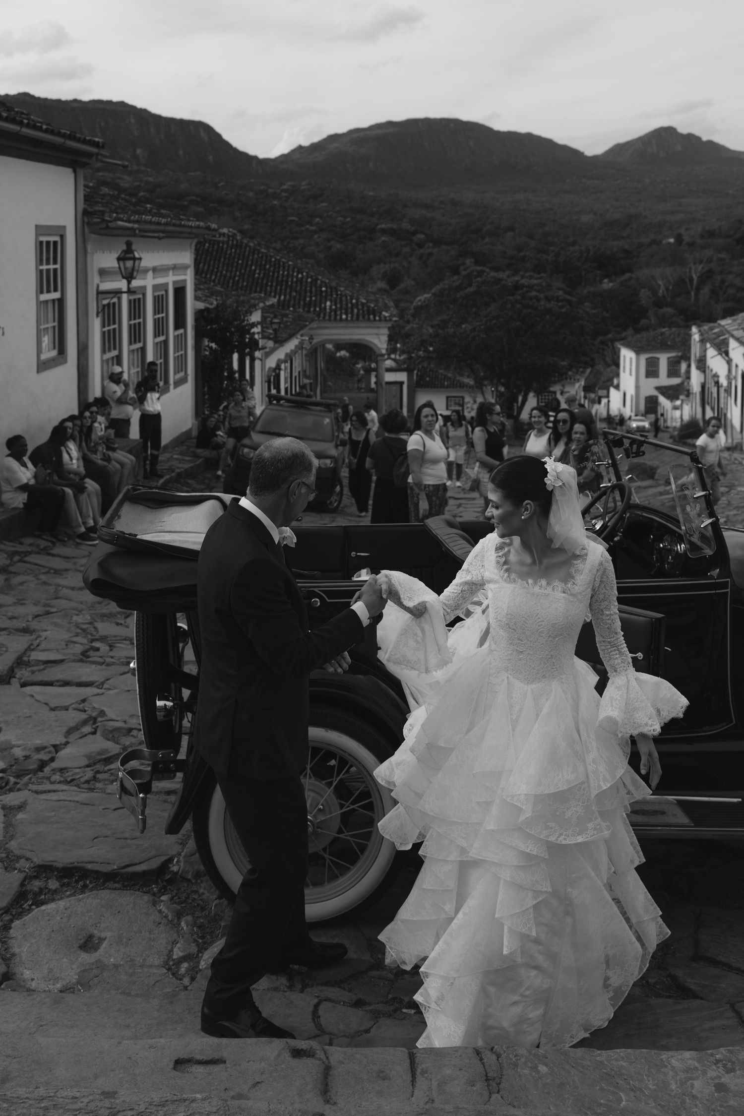 Black and white wedding photo of bride and groom holding hands beside vintage car on cobblestone street in historic town.