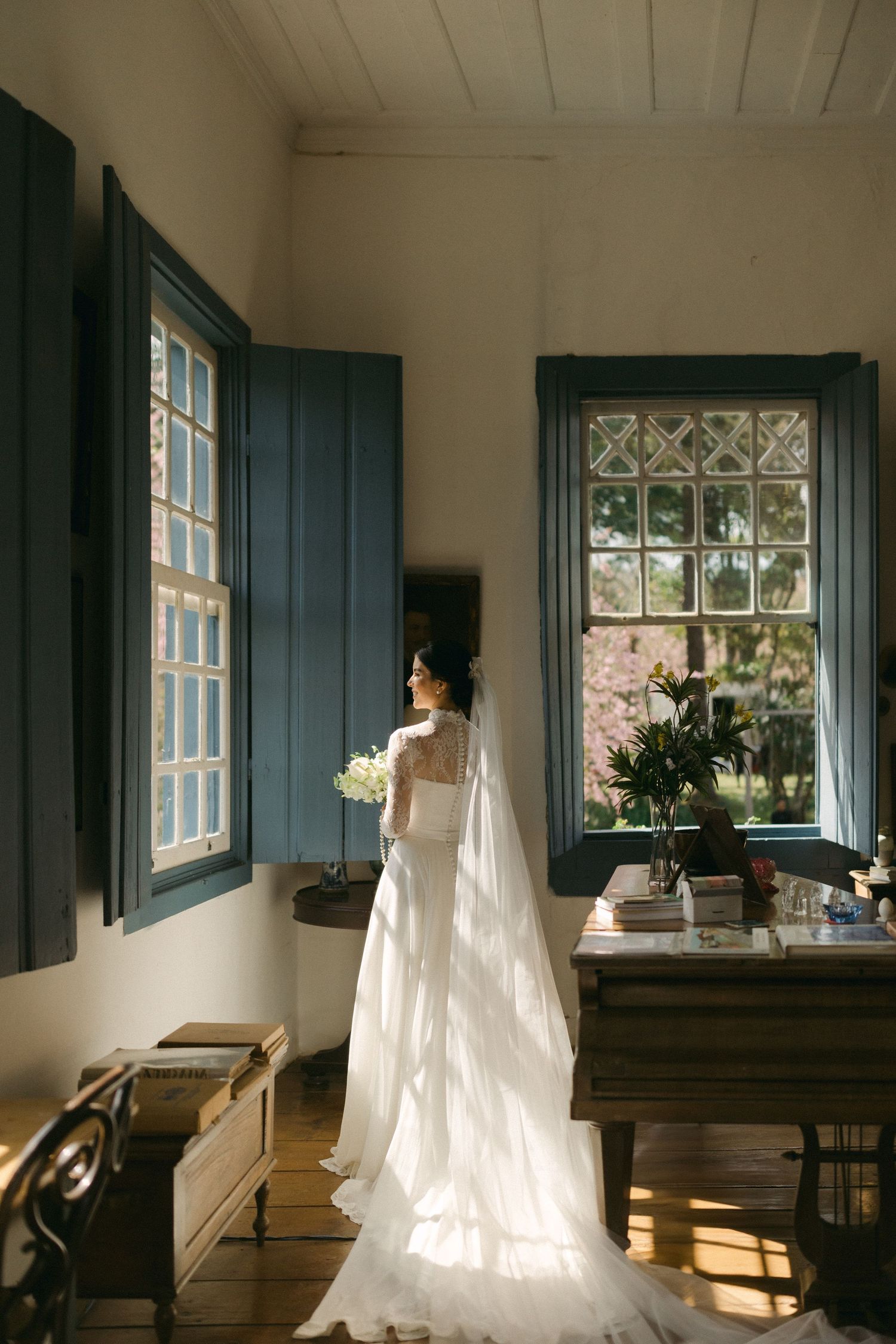 A long white wedding dress and veil catching sunlight through vintage windows in a rustic room with green shutters.