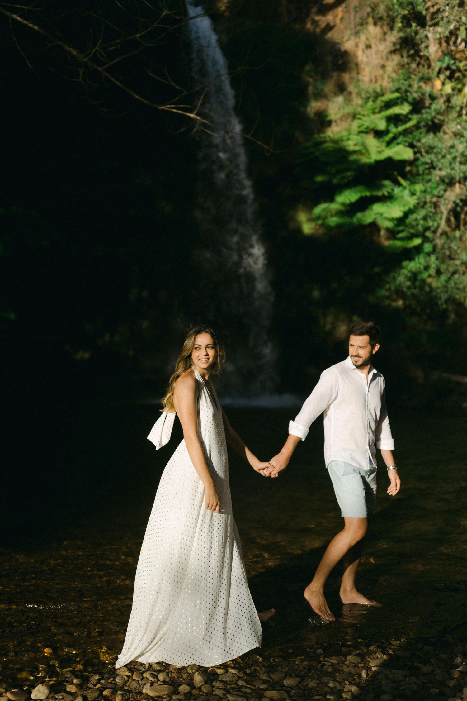 A couple holds hands while exploring near a dramatic waterfall in white attire.