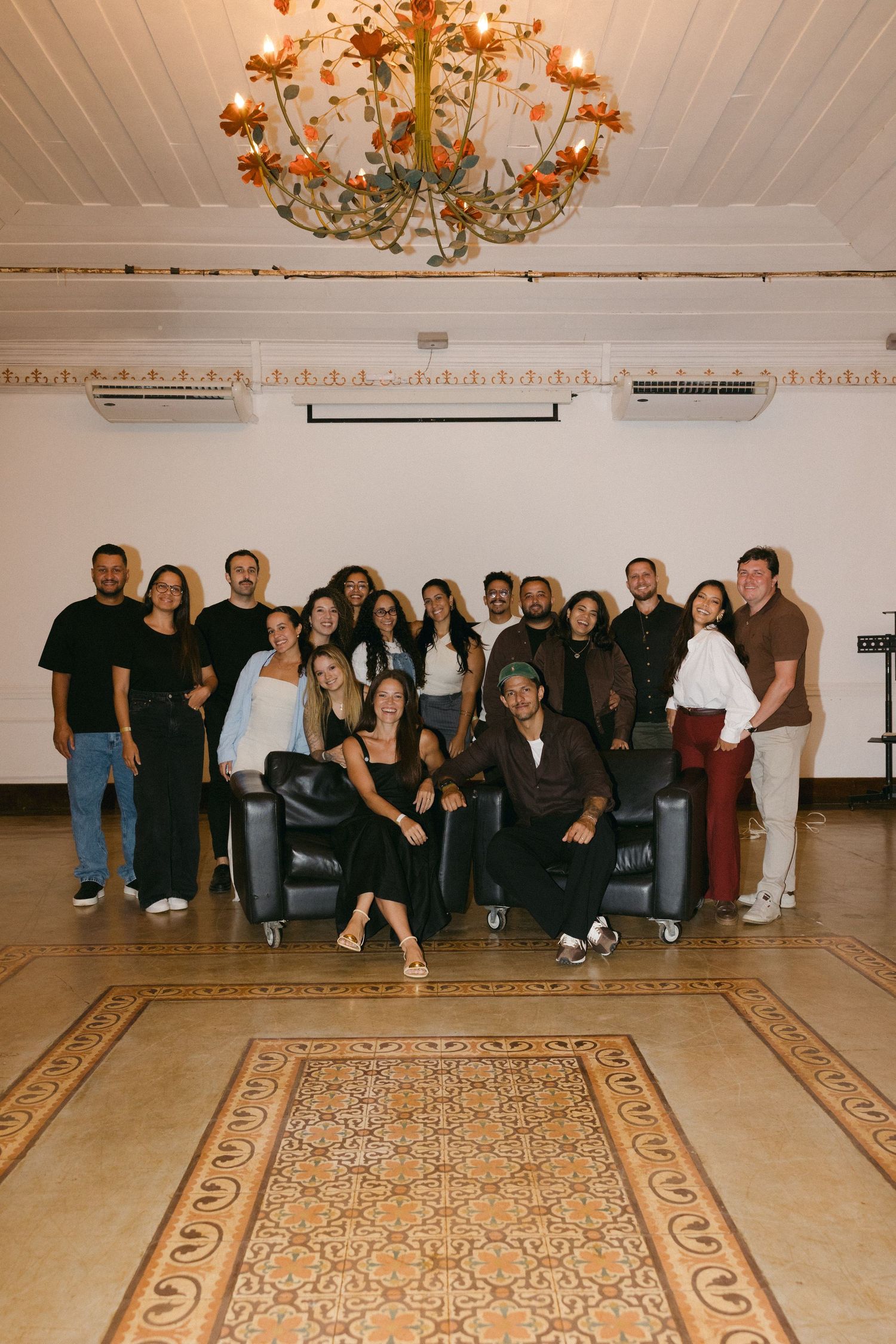 Group of young adults posing together in an elegant room with ornate chandelier and decorative tile floor.