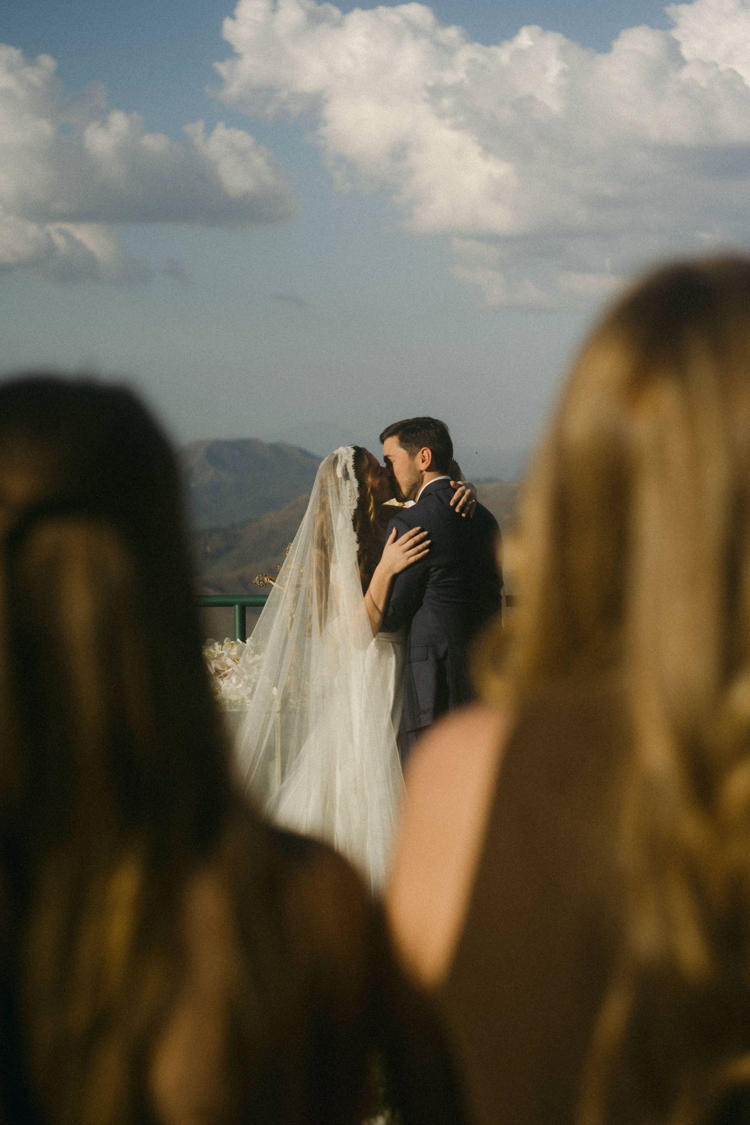 Bride and groom share first kiss at outdoor wedding ceremony with mountain backdrop and cloudy blue sky.