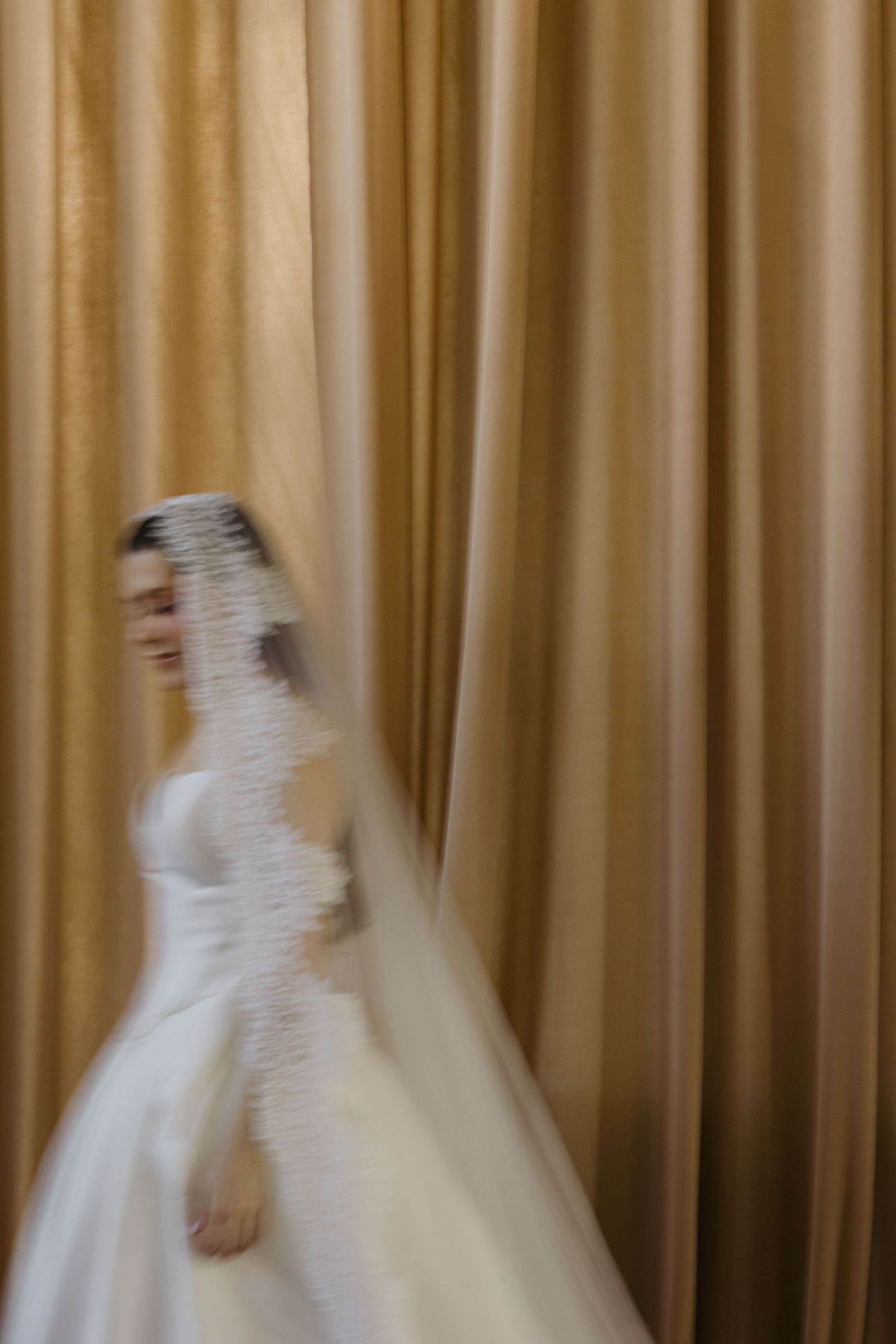 A blurred bride in a white wedding gown and veil poses gracefully against golden draped curtains.