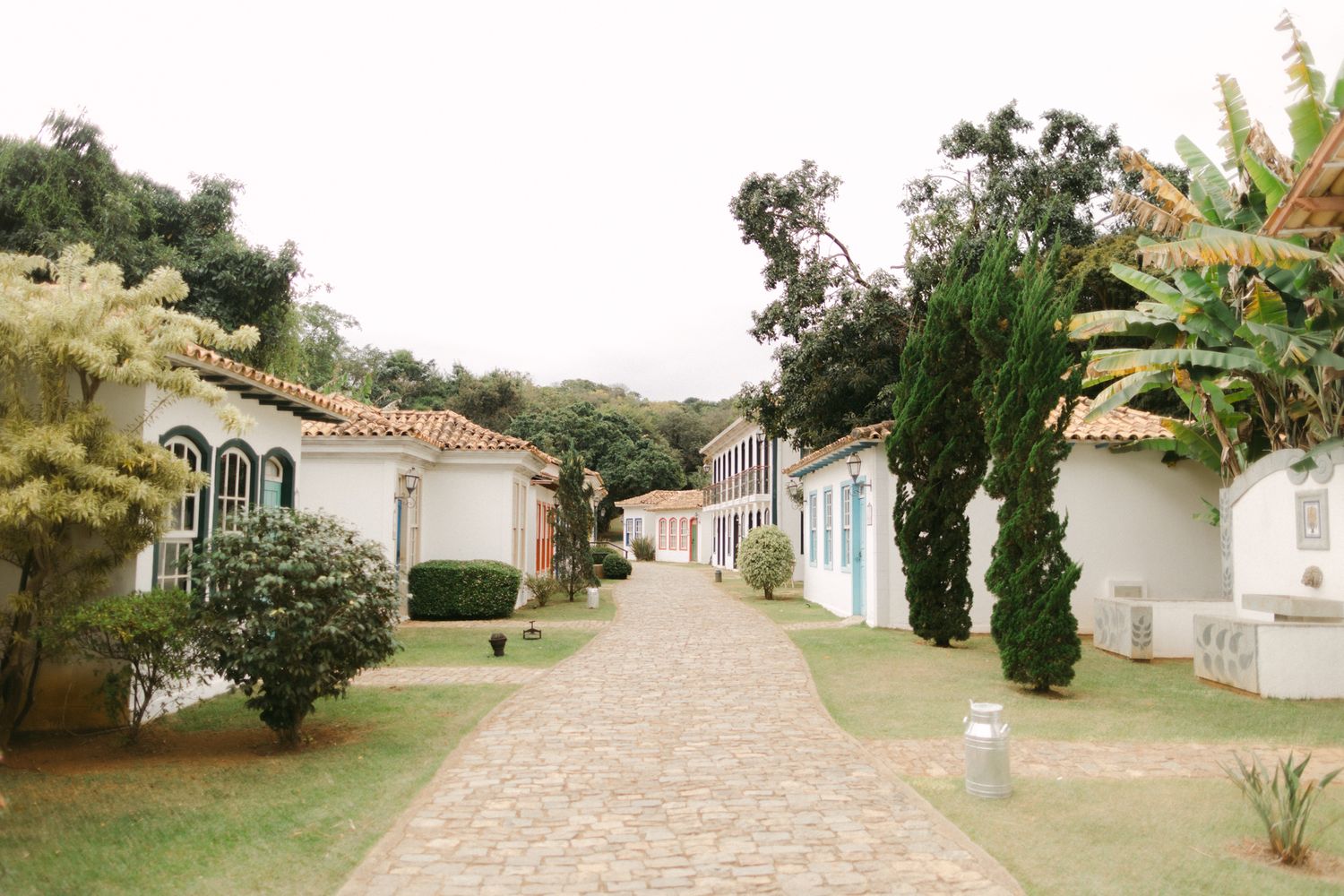 A row of white bungalow-style houses with terracotta roofs line a winding path surrounded by lush tropical vegetation.