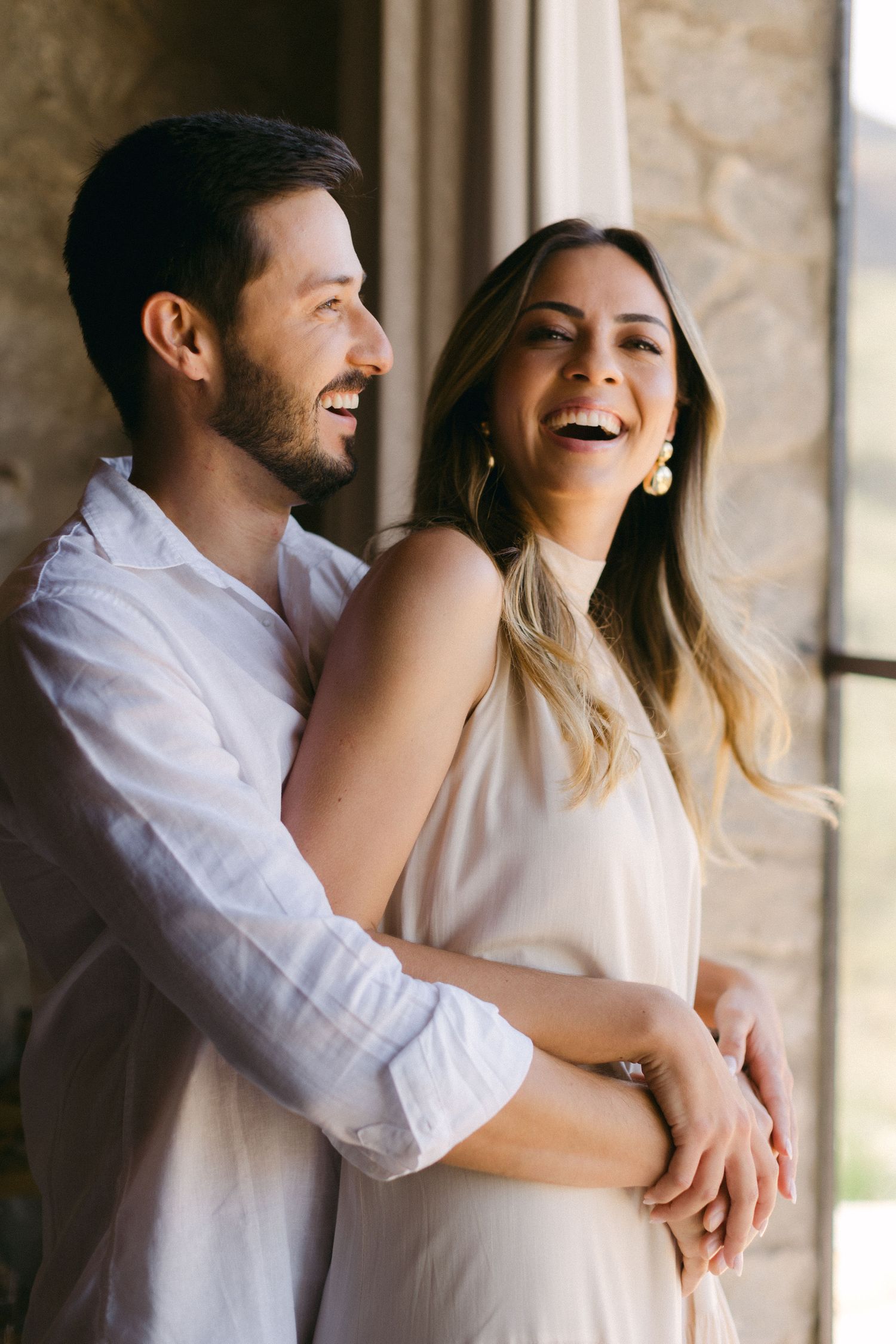A joyful indoor portrait of a couple laughing and embracing in natural window light.