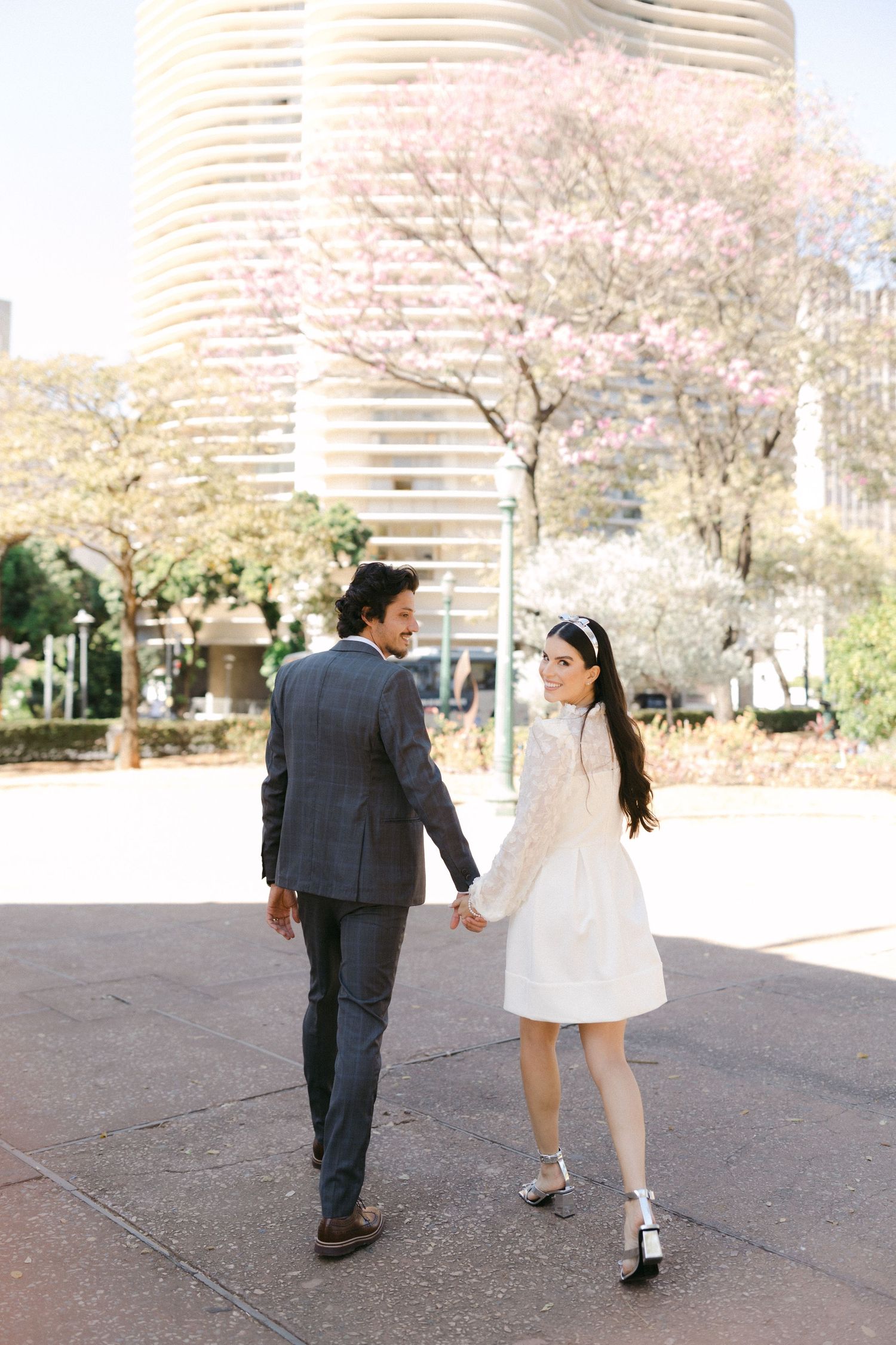 A couple holding hands while walking near cherry blossoms and modern architecture.