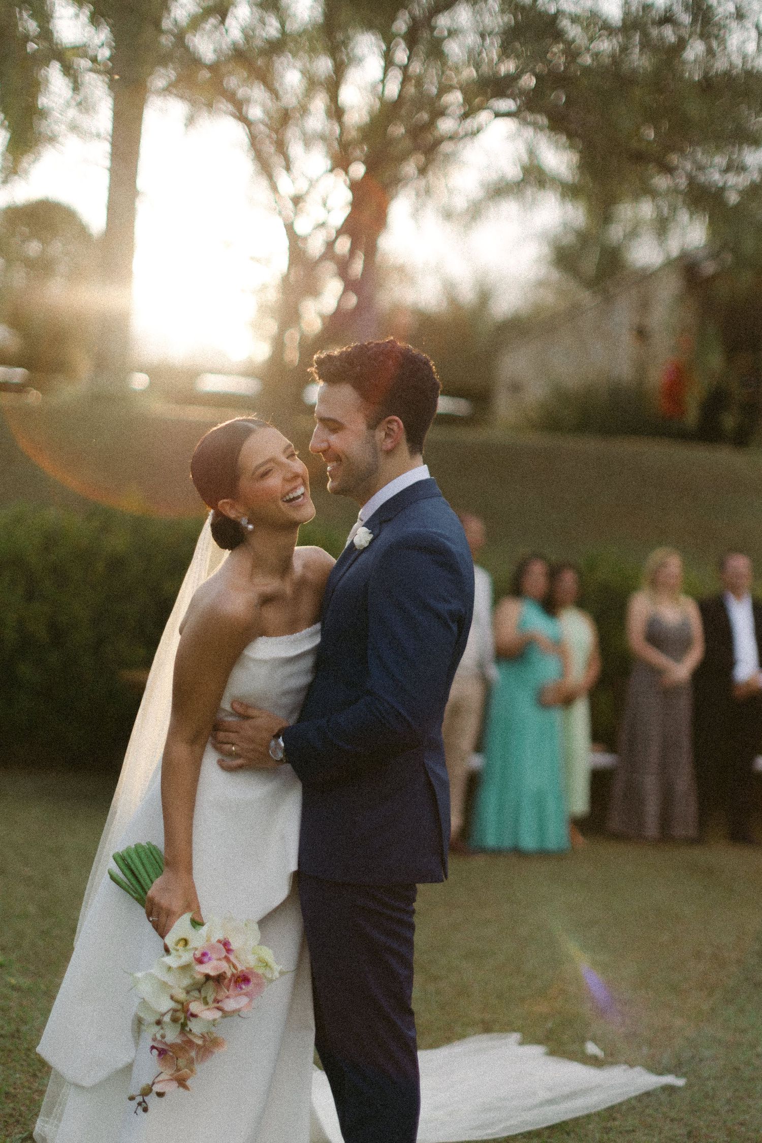 A joyful wedding moment captured at sunset with guests in mint green and gray dresses watching in the background.