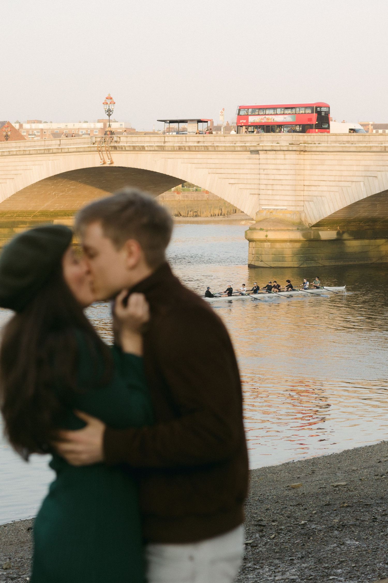 Couple embraces near historic bridge with red double-decker bus crossing in background.