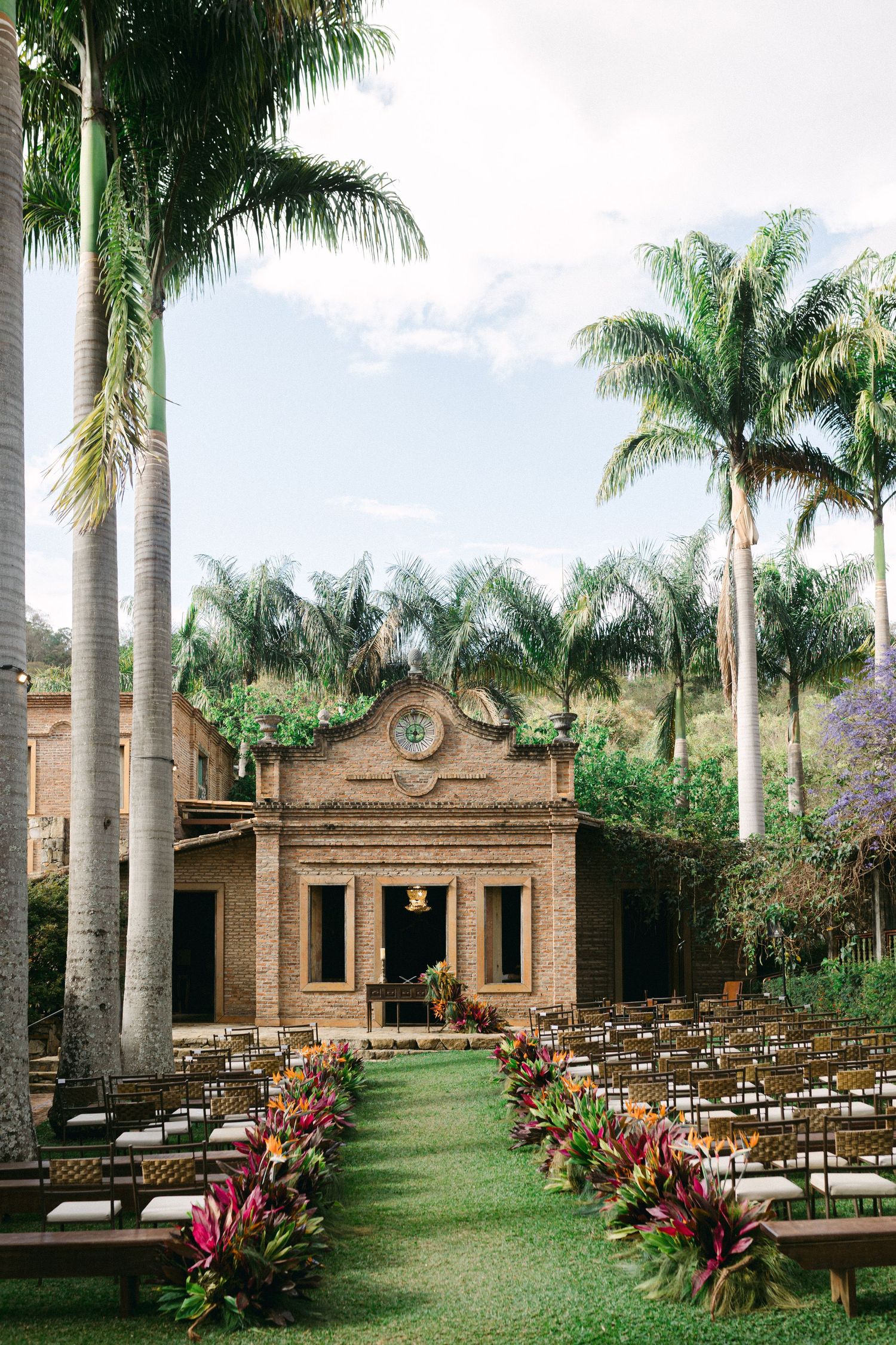Outdoor wedding ceremony setup with tropical palm trees, colorful floral aisle, wooden chairs, and rustic brick chapel.