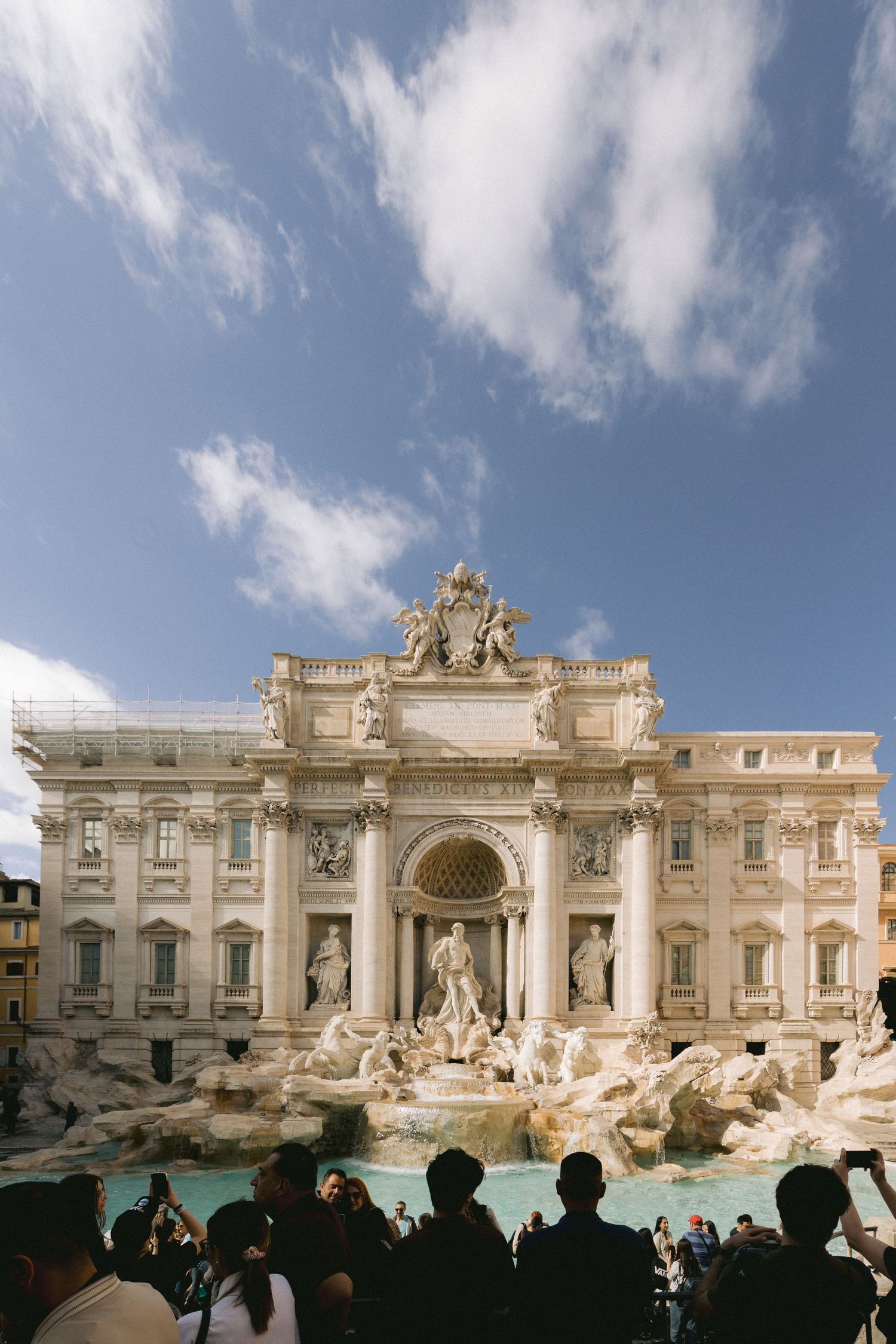 A panoramic view of the iconic Trevi Fountain in Rome with ornate Baroque architecture and tourists silhouetted against the water.