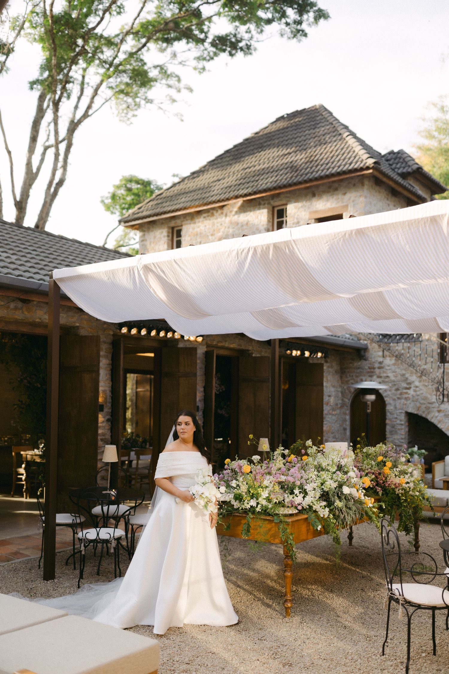 White canopy shades an outdoor patio area with rustic stone walls and floral arrangements at an elegant venue.