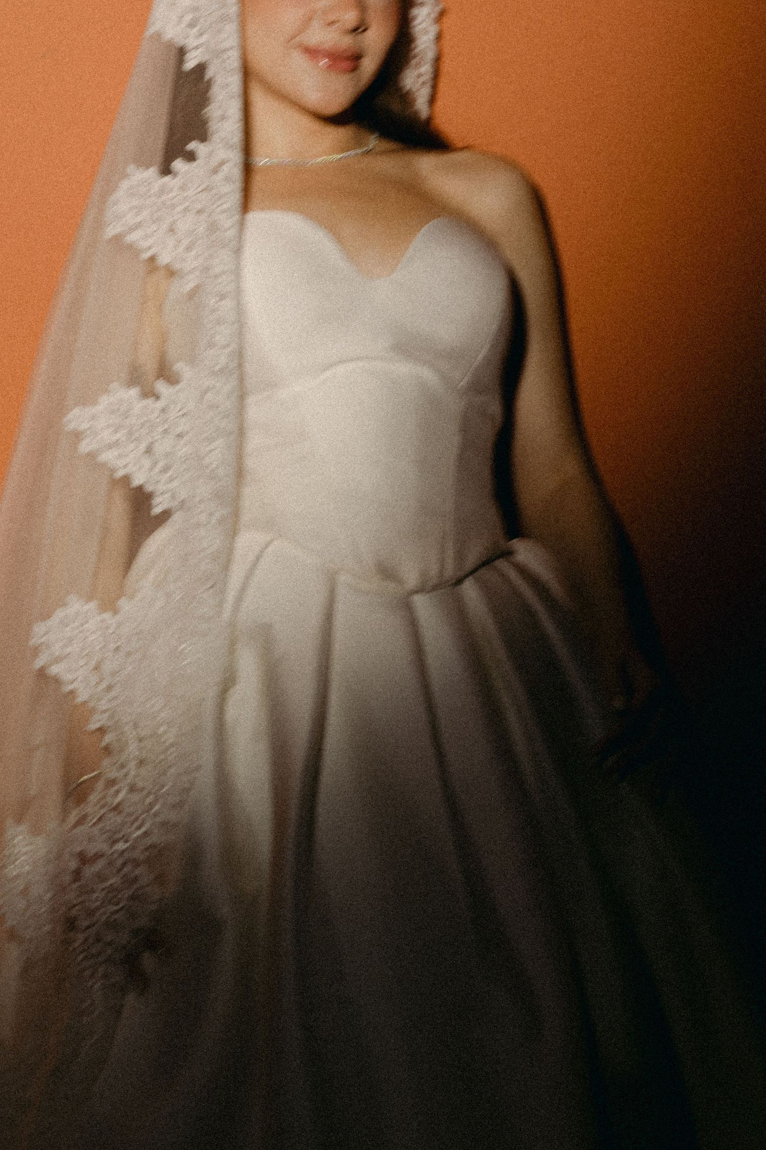 Bride in strapless white ball gown with lace-trimmed veil against warm orange background.