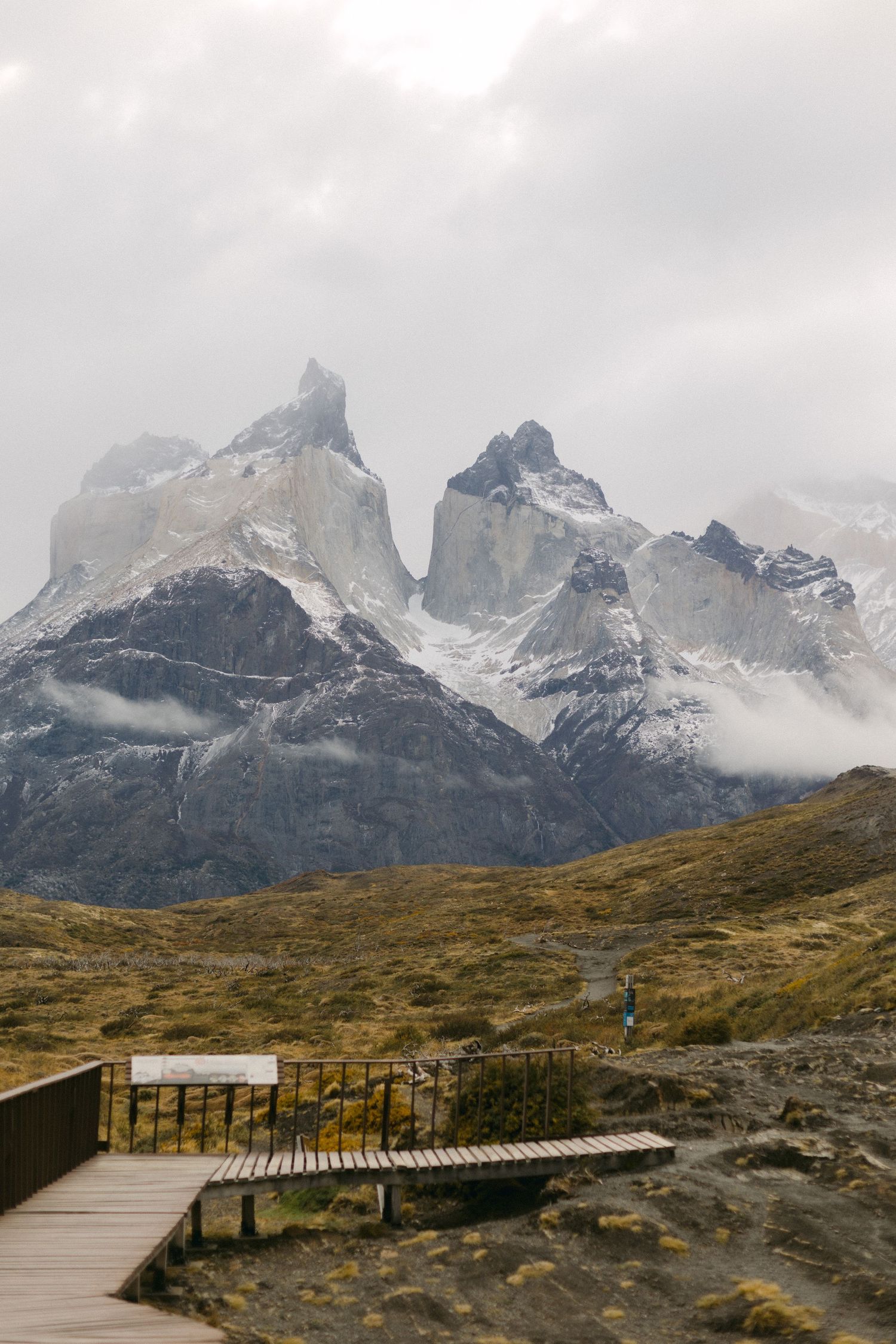 Wooden boardwalk trail winding through Torres del Paine National Park with dramatic mountain peaks shrouded in clouds.