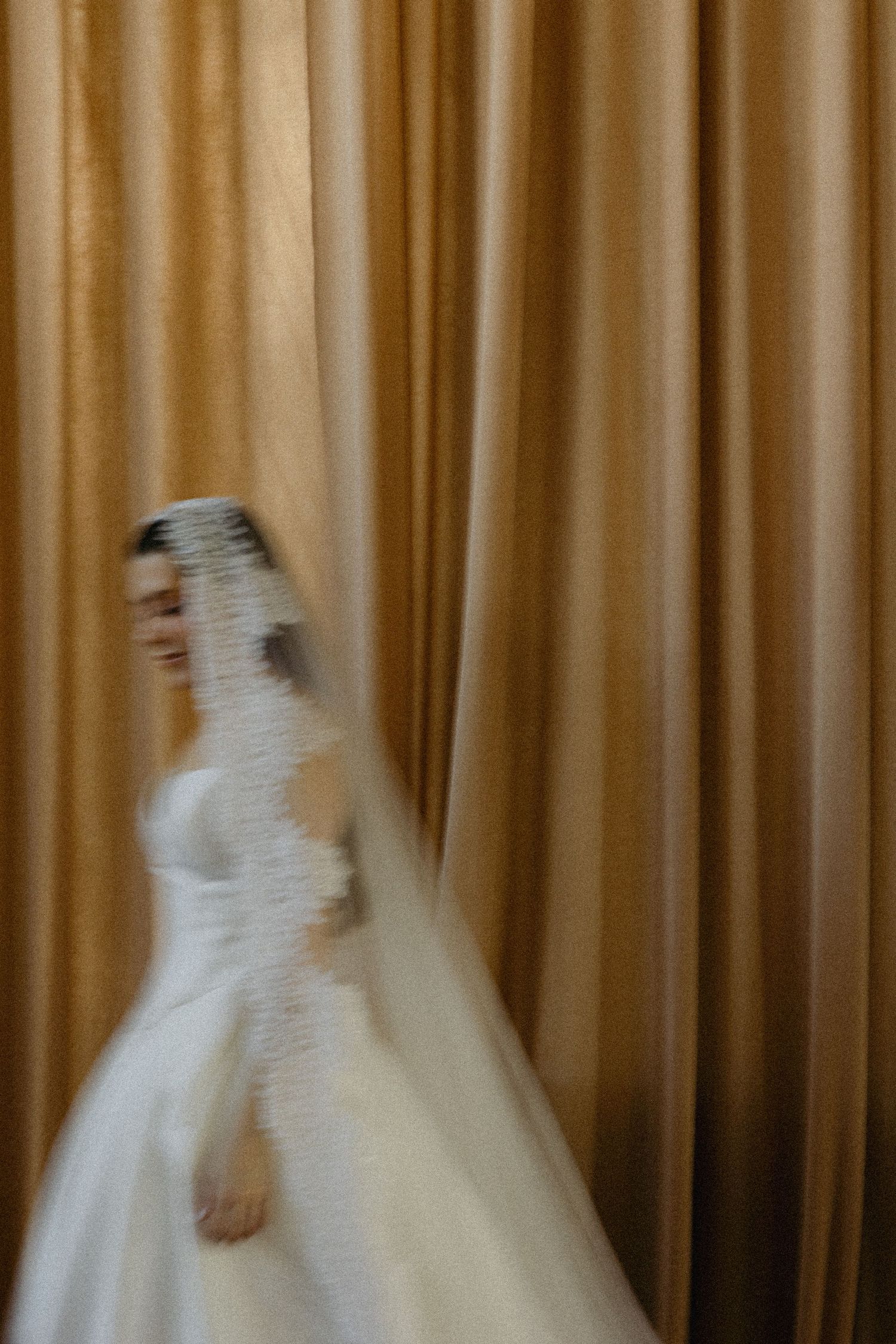 A blurred bride in a white wedding gown and veil poses gracefully against golden draped curtains.