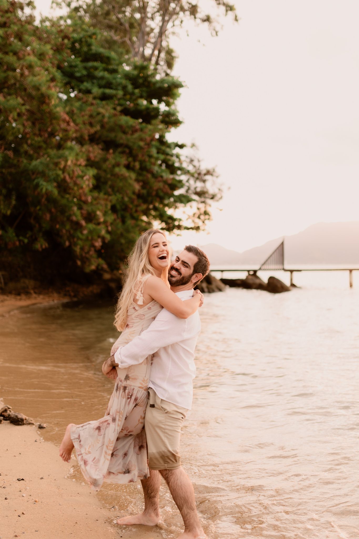 A joyful couple embraces on a beach at sunset with boats visible in the background.