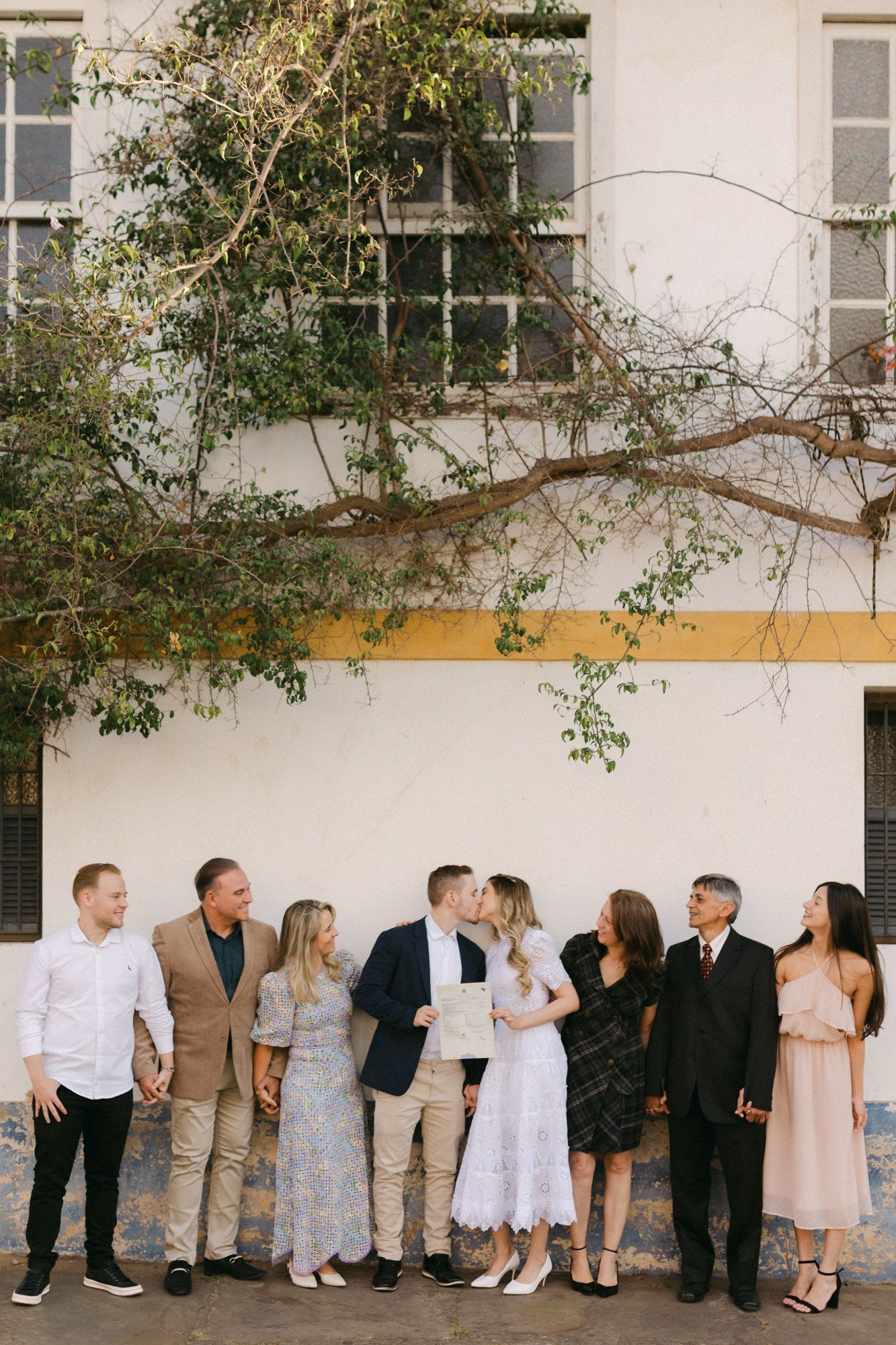 Wedding party lined up against white building wall with overhanging tree branches.