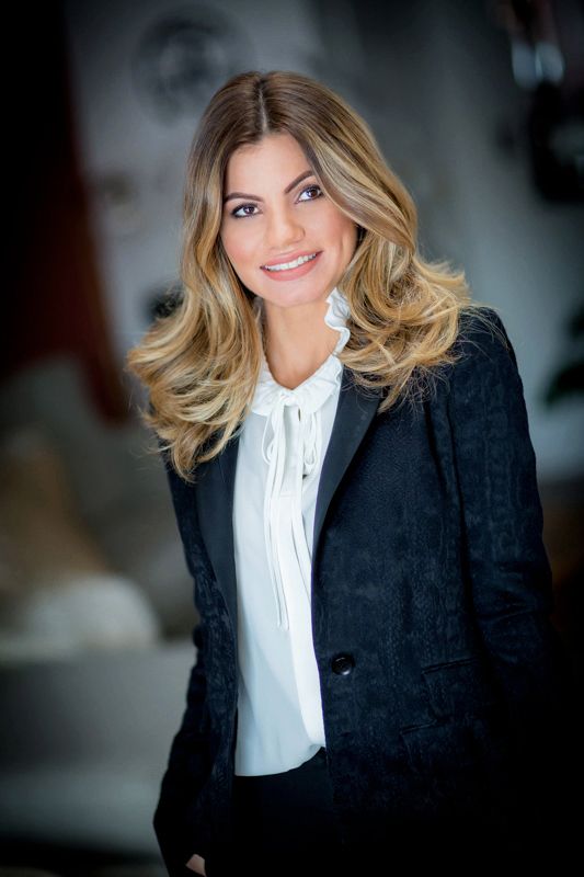 Professional headshot featuring blonde hair, white blouse and black blazer against blurred background.