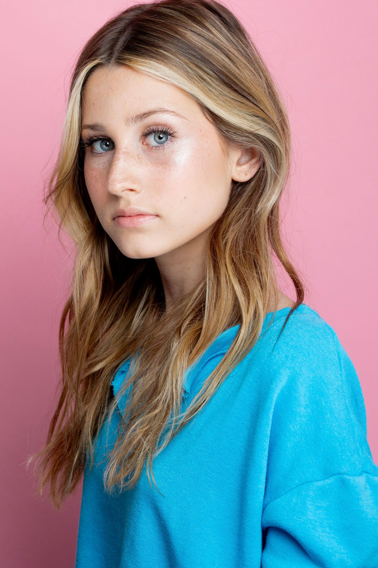 Portrait against pink backdrop shows blonde hair and bright turquoise sweater in studio lighting.