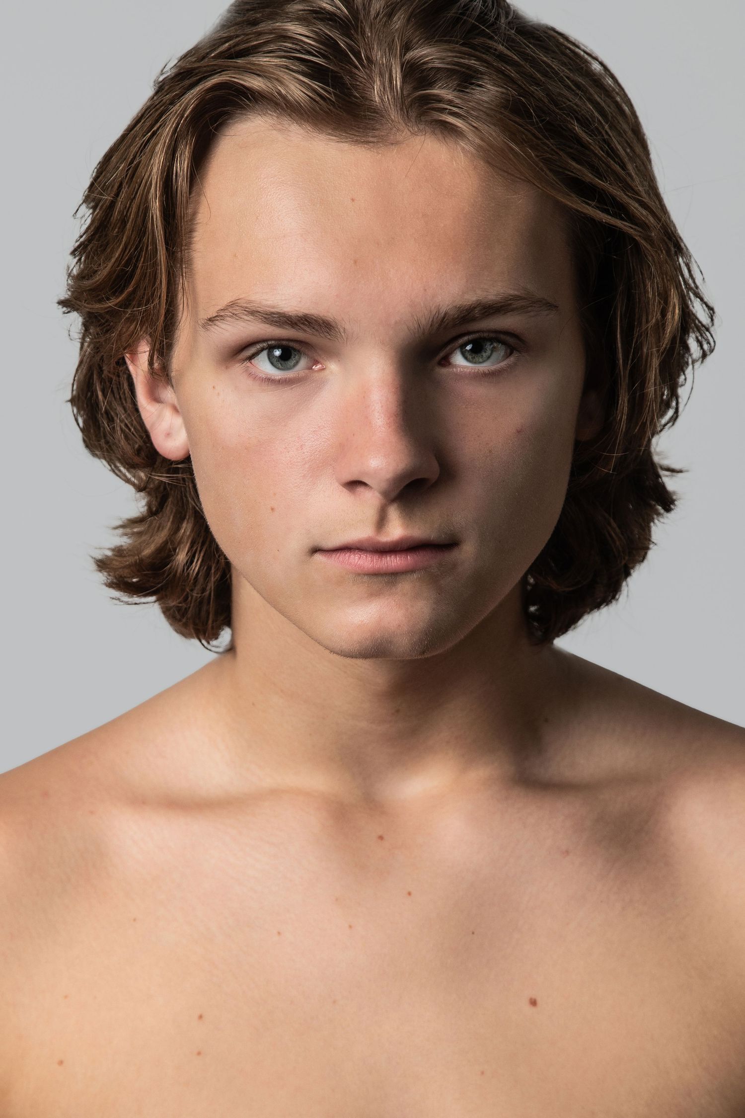 Headshot portrait of young person with shoulder-length brown hair against light background.