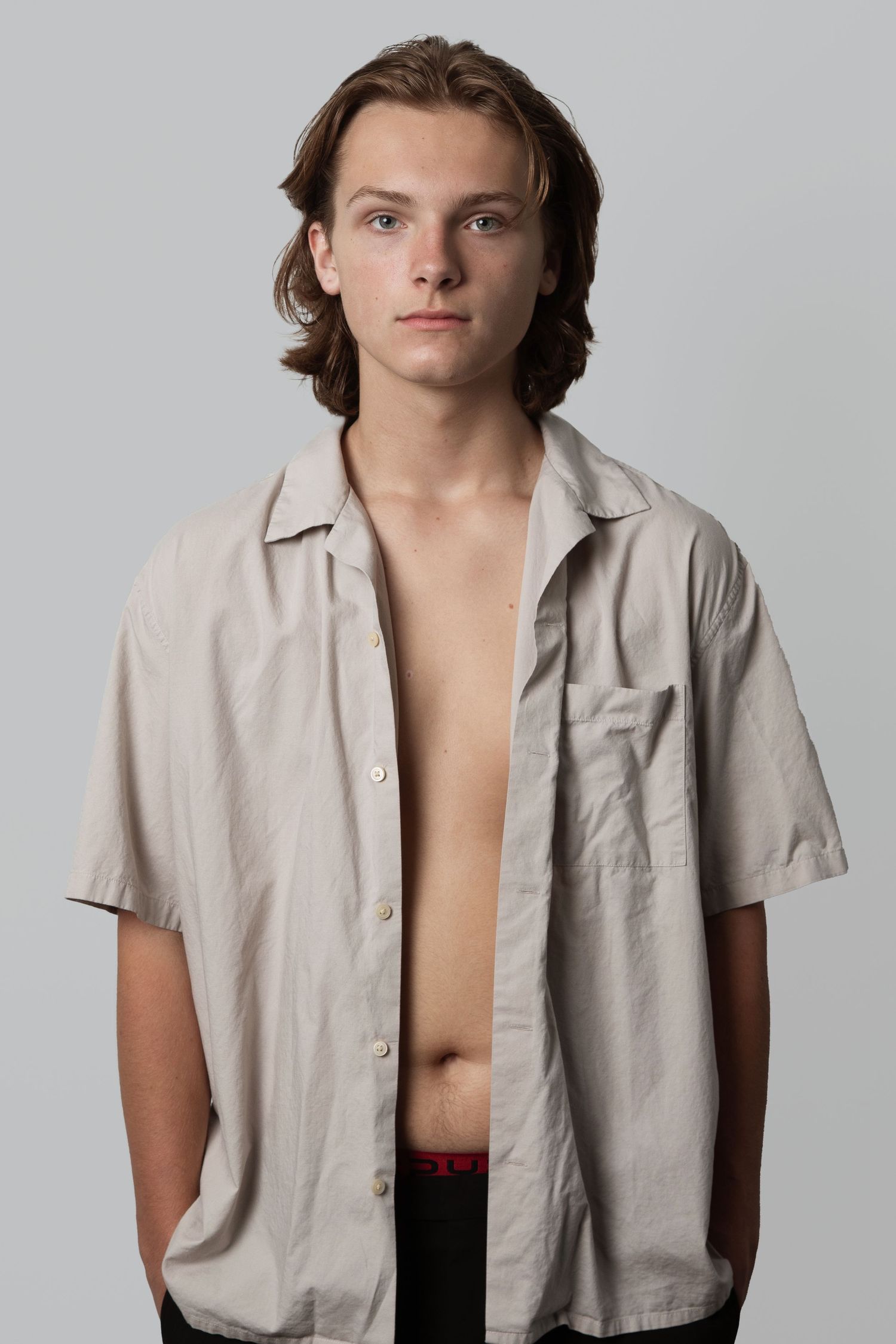 Young male model in unbuttoned beige short-sleeve shirt against gray background.