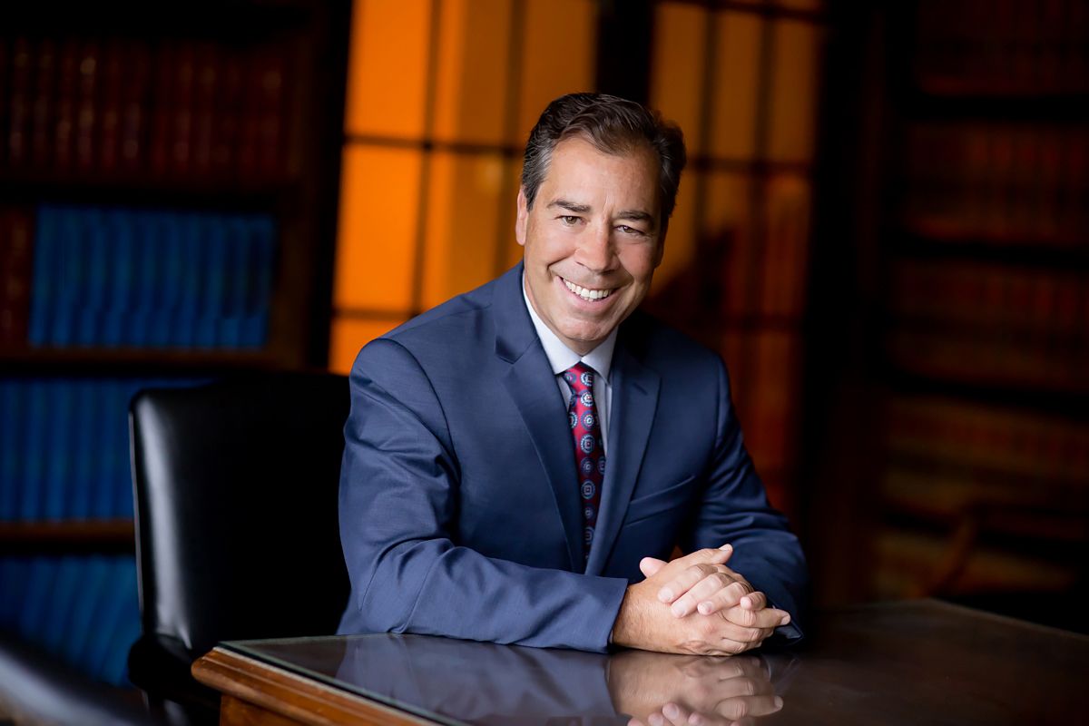 Executive portrait of person in navy suit seated at desk with warm lighting and mahogany background.