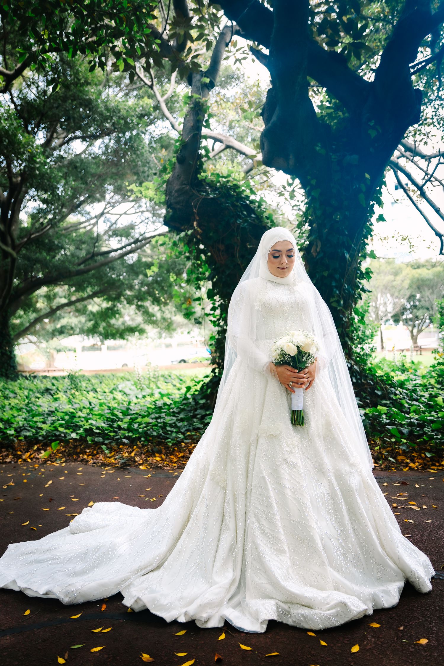 Muslim bride in white lace gown and hijab holding bouquet in lush green park setting. Wollongong Wedding Photographer