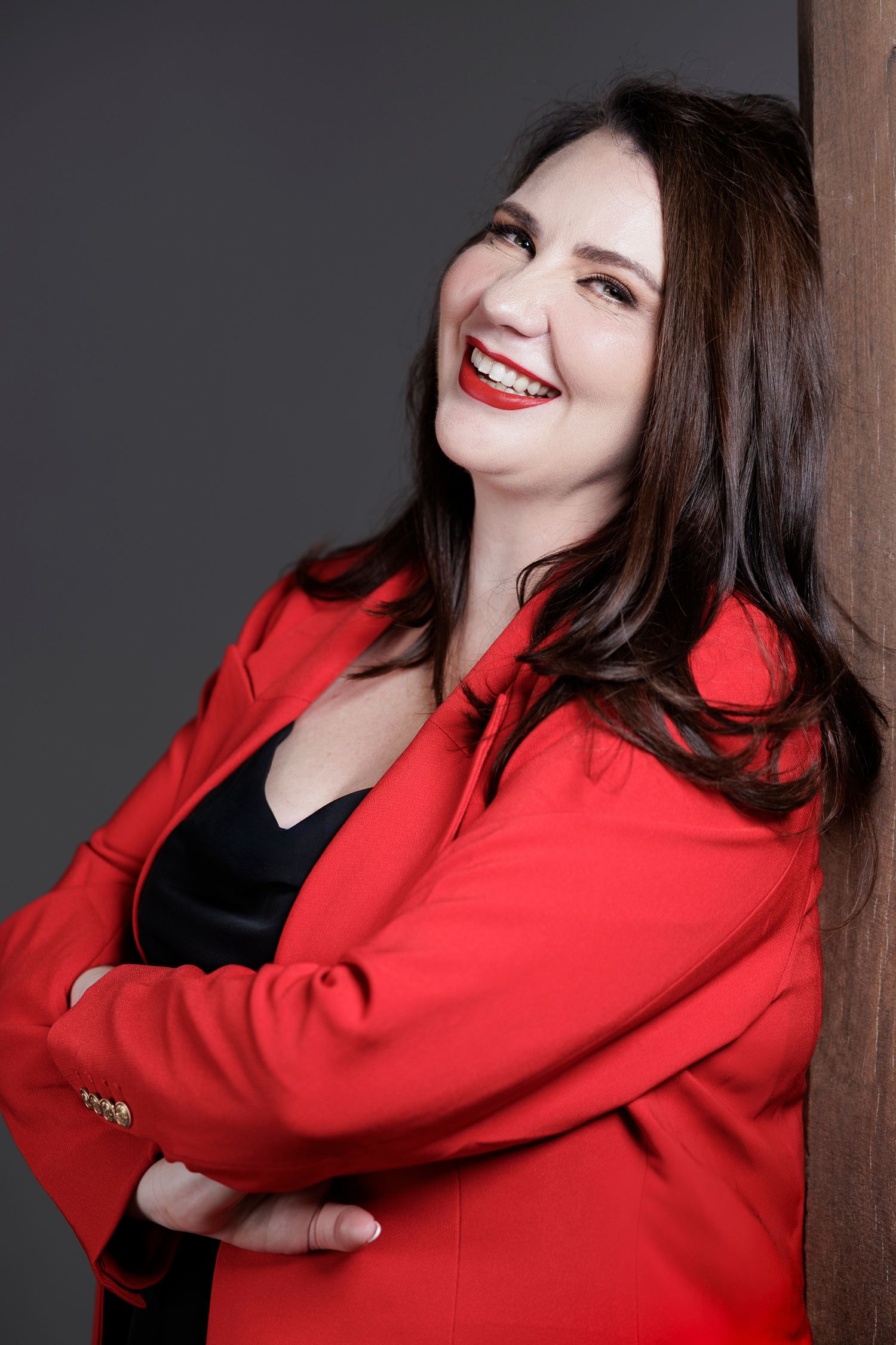 Wollongong Corporate Branding Photographer. A smiling woman in a red blazer with red lipstick leans against a wall, arms crossed.