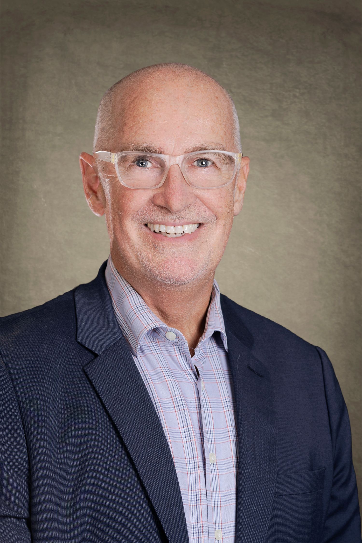 Professional headshot of a smiling bald man wearing clear glasses and a navy blazer over a plaid shirt. Wollongong Headshot Photographer