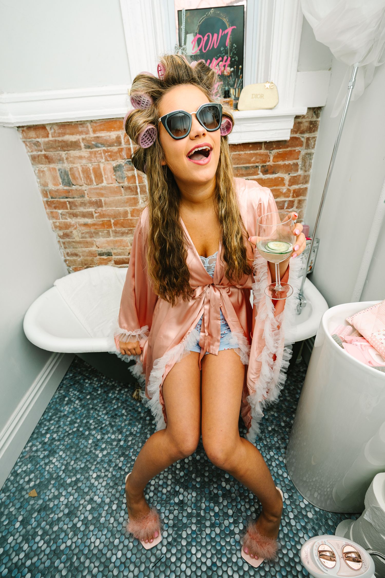 Wollongong Portrait Photographer. Woman in pink robe, hair rollers, and sunglasses sitting in bathroom holding a champagne glass.