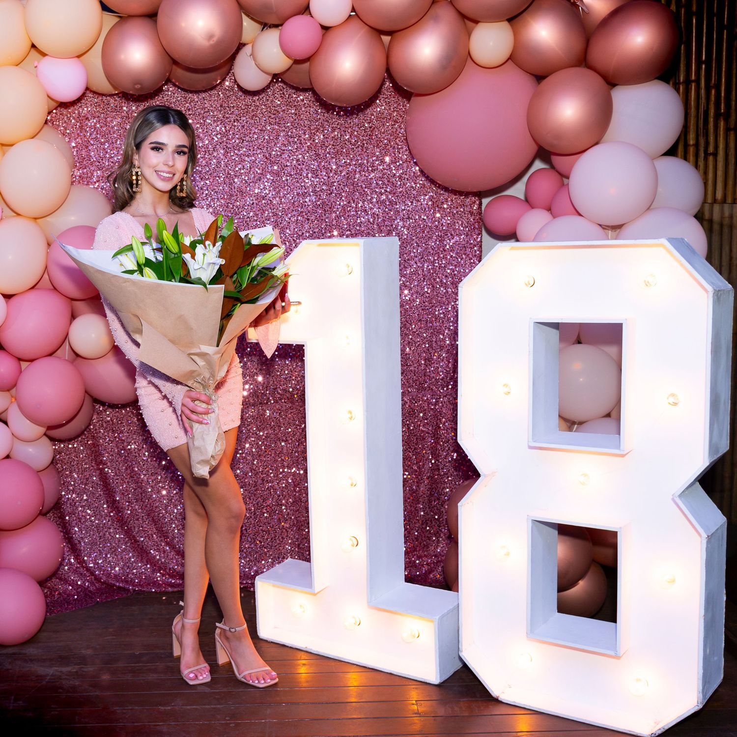Young woman holding flowers poses beside illuminated number 18 sign at a pink balloon-decorated 18th birthday. Sydney Event Photographer