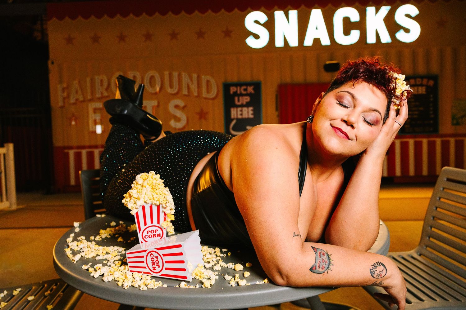 Wollongong Portrait Photographer. Woman posing with popcorn at a carnival snack bar, wearing a black dress with a tattoo on her arm.