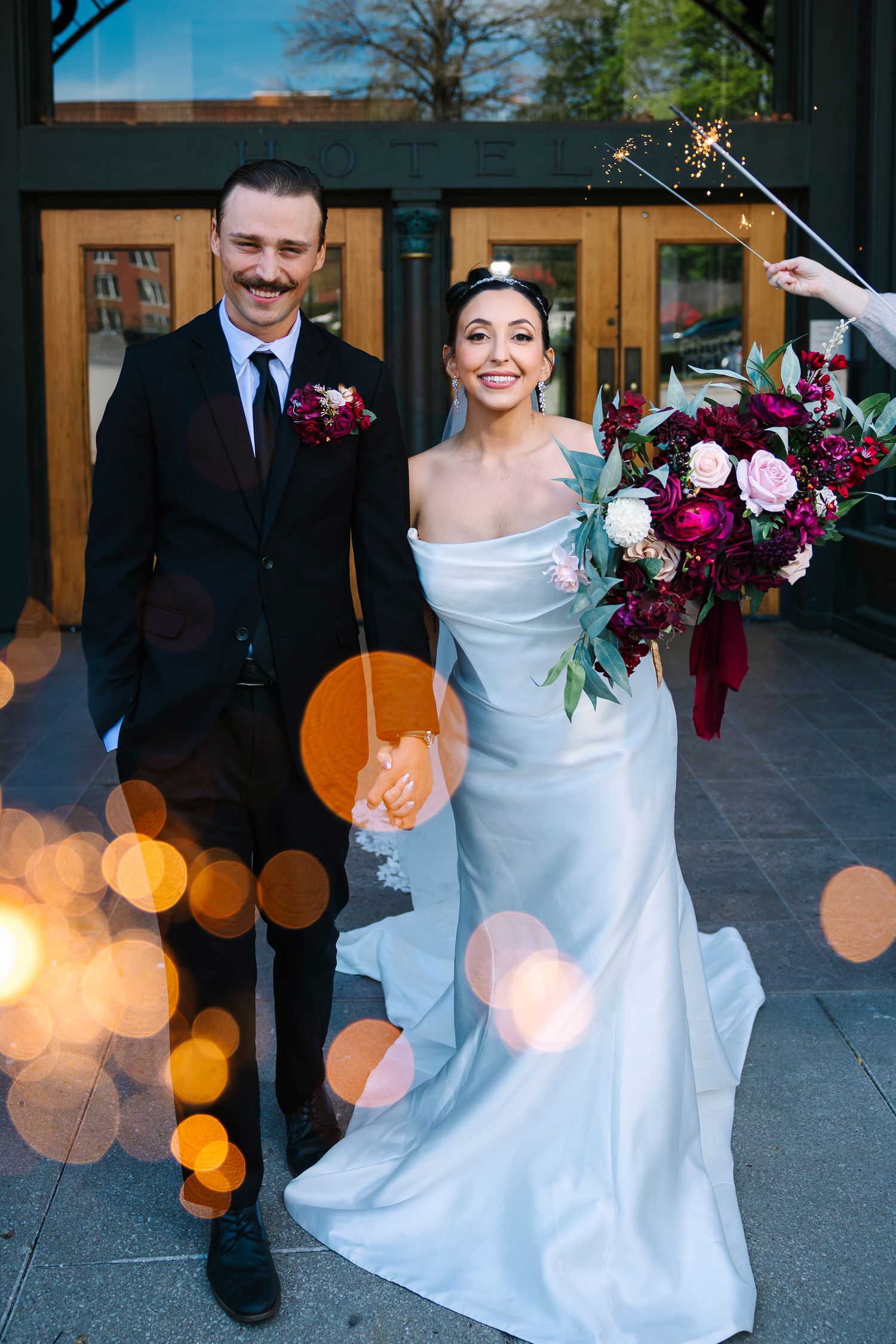 Bride in wedding gown holds burgundy bouquet beside groom in black suit, sparklers glowing at wedding exit. Wollongong Wedding Photographer