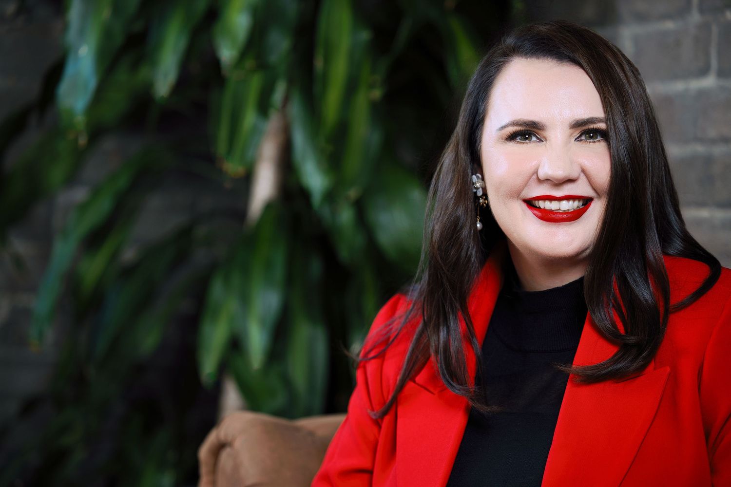 Professional woman in red blazer smiling confidently, posed against lush green foliage background. Wollongong personal branding photography.